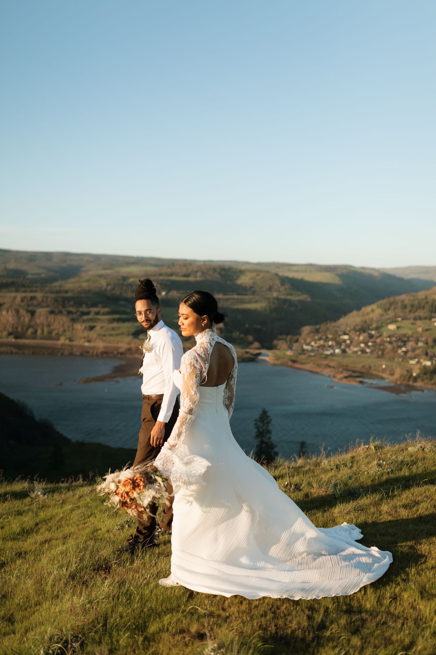 Bride in lace wedding gown holding floral bouquet stands with groom overlooking scenic river valley landscape.
