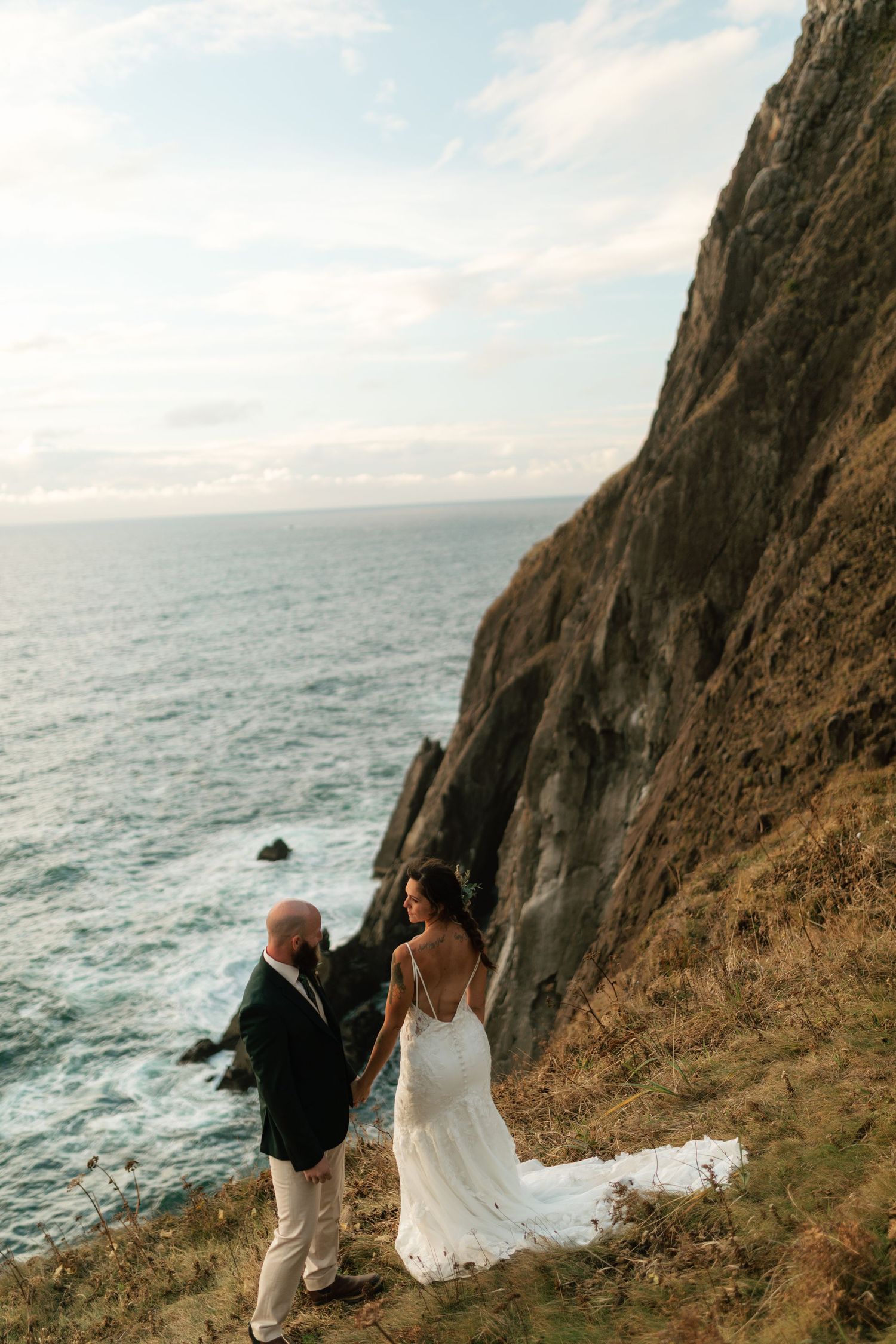 Bride and groom hold hands on a dramatic coastal cliff overlooking the ocean in wedding attire.