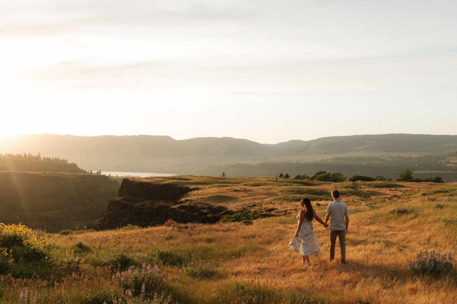 Couple walking hand-in-hand through golden meadow with sweeping river valley sunset views.