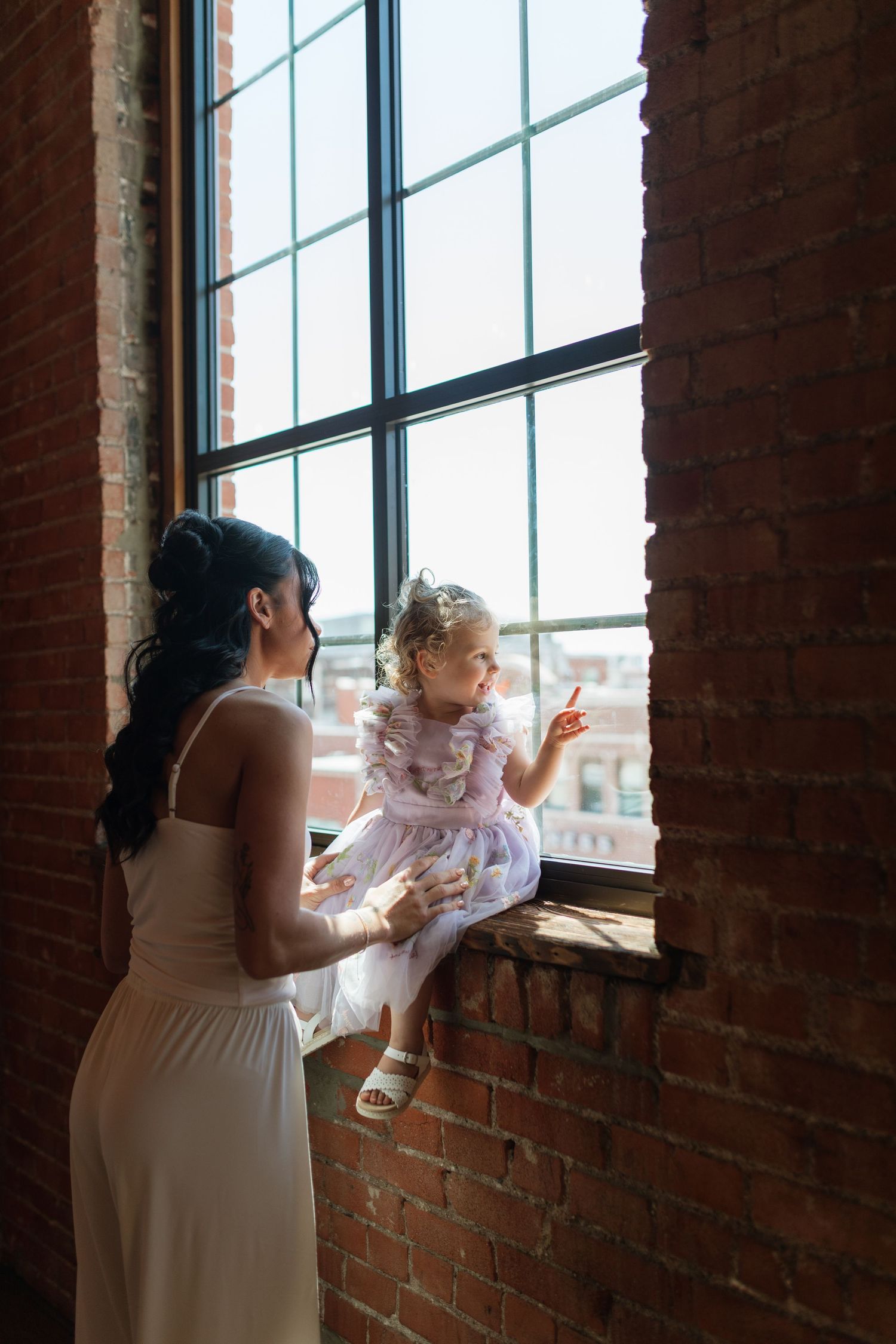 Bridesmaid holding a young flower girl sitting on a windowsill in a brick-walled venue.