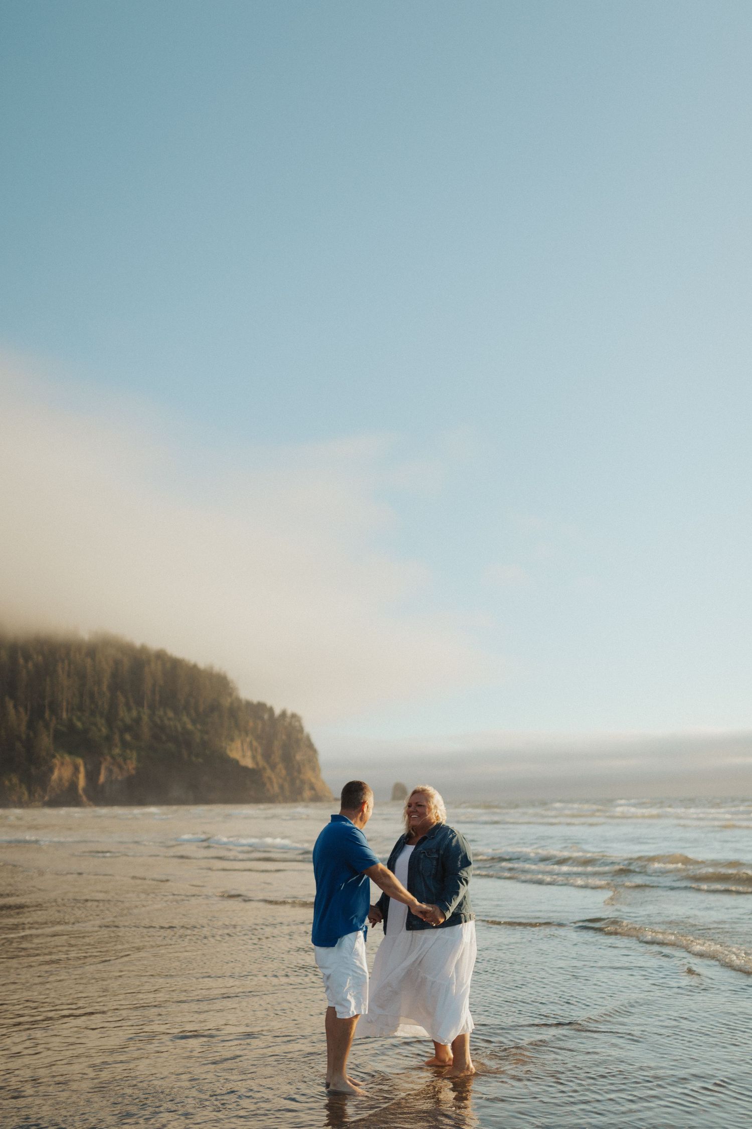 Mature couple holding hands in ocean water on a scenic beach with foggy cliffs in background.