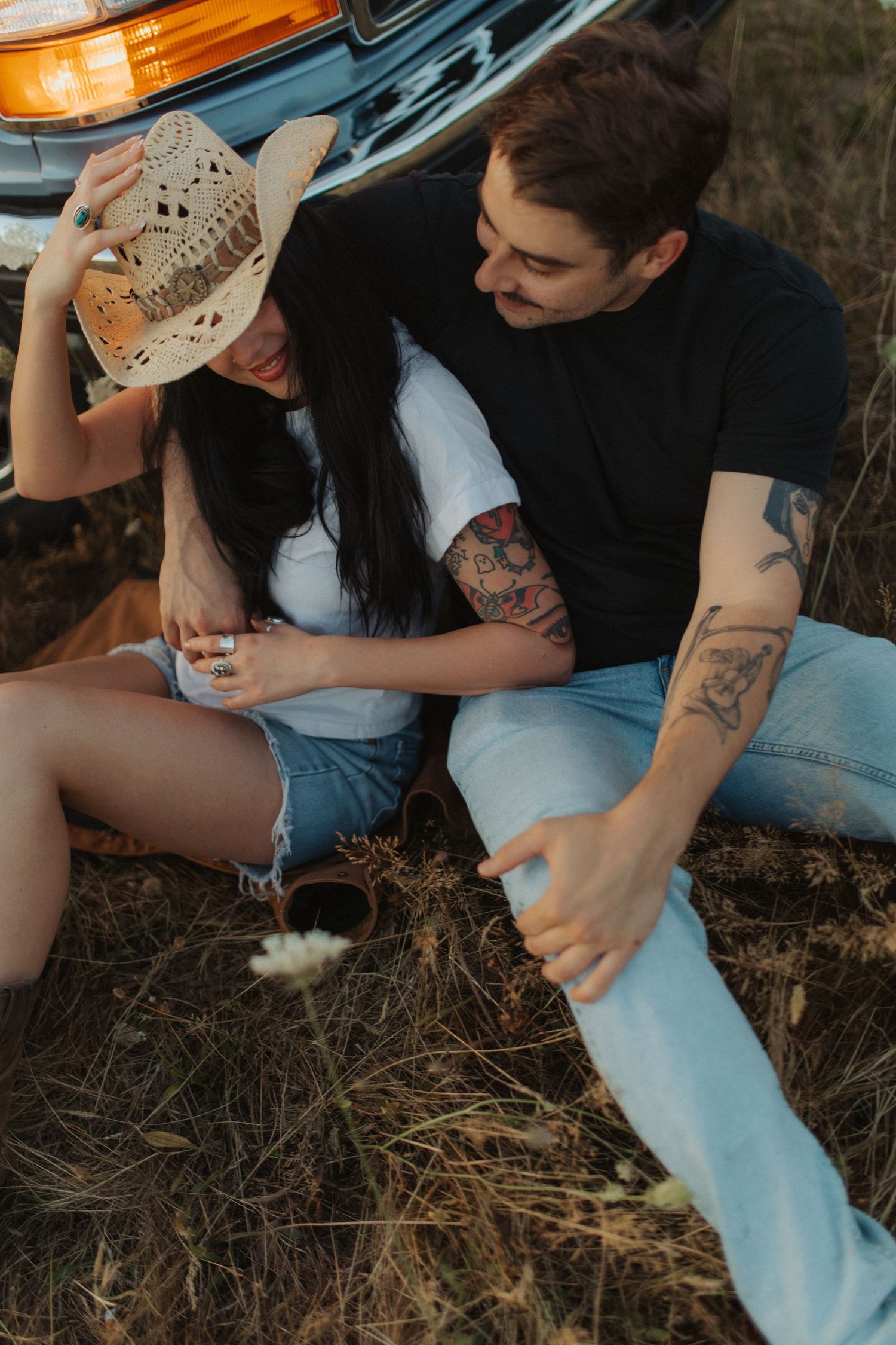 A tattooed couple sits together in a field leaning against a vintage truck at sunset.