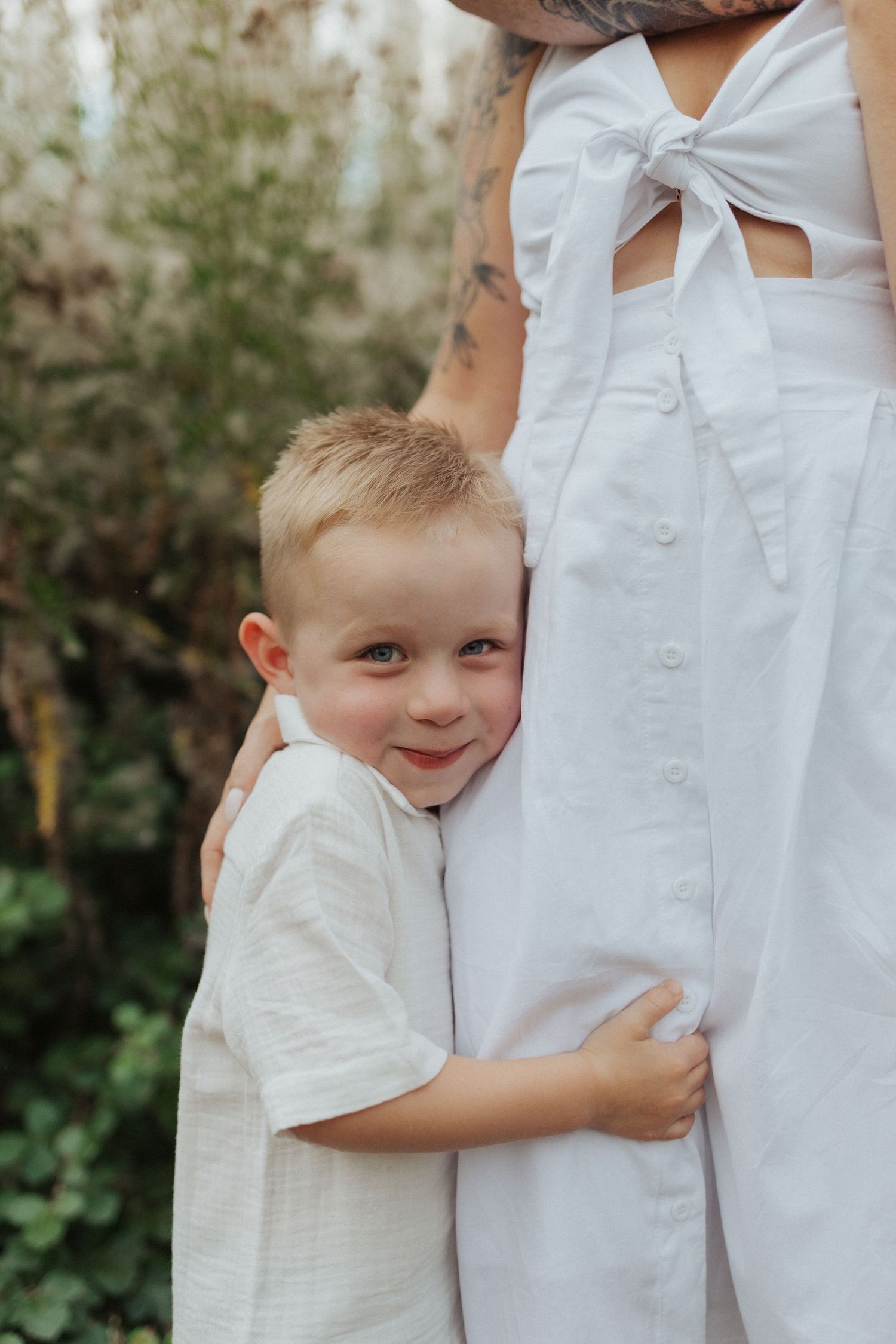 Adorable blonde toddler boy hugging his mothers white dress outdoors in a garden setting.
