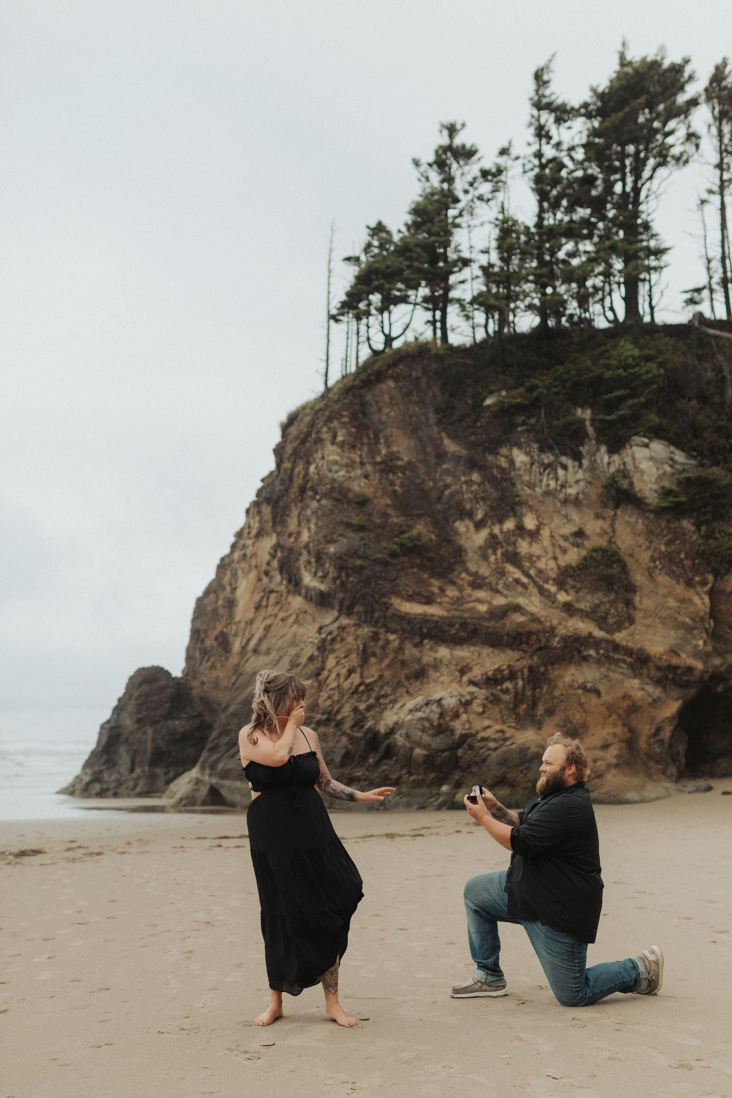 Man on one knee proposing to woman in black dress on a misty beach with rocky cliffs.