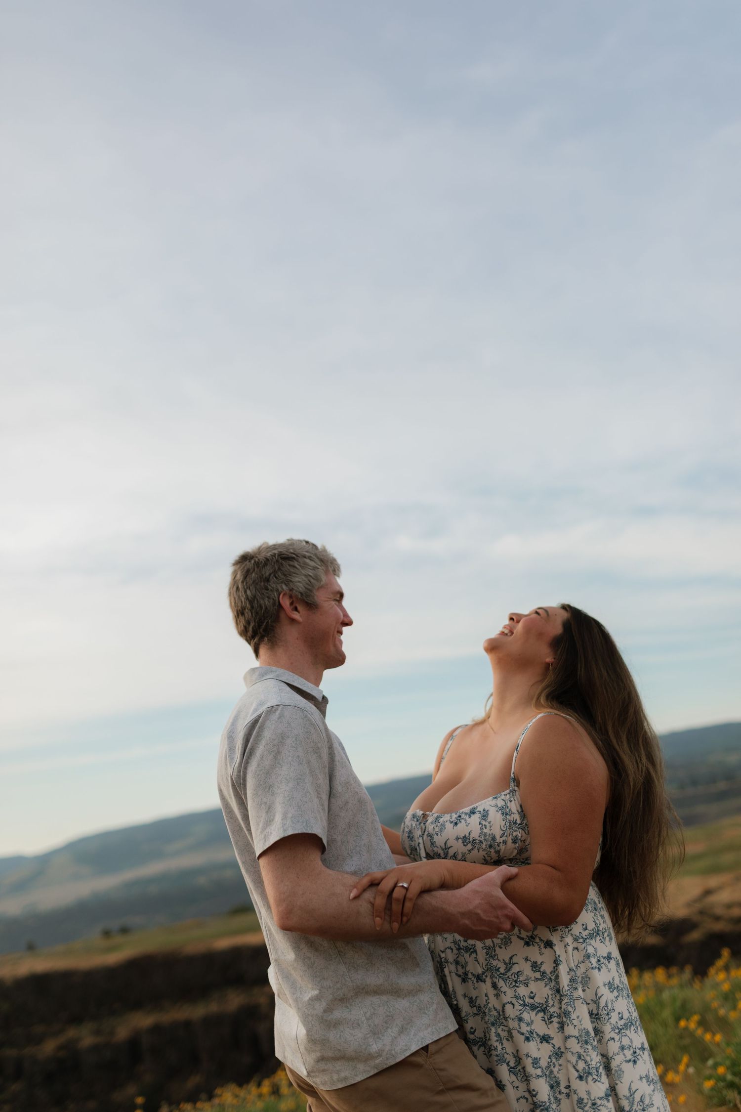 Couple holding hands and laughing together in a golden field with rolling hills in the background.