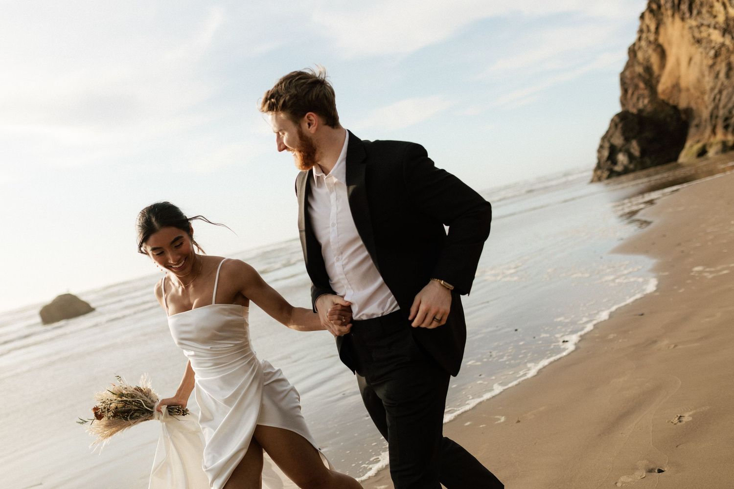Joyful newlyweds run hand-in-hand along a sandy beach in wedding attire with ocean waves behind them.