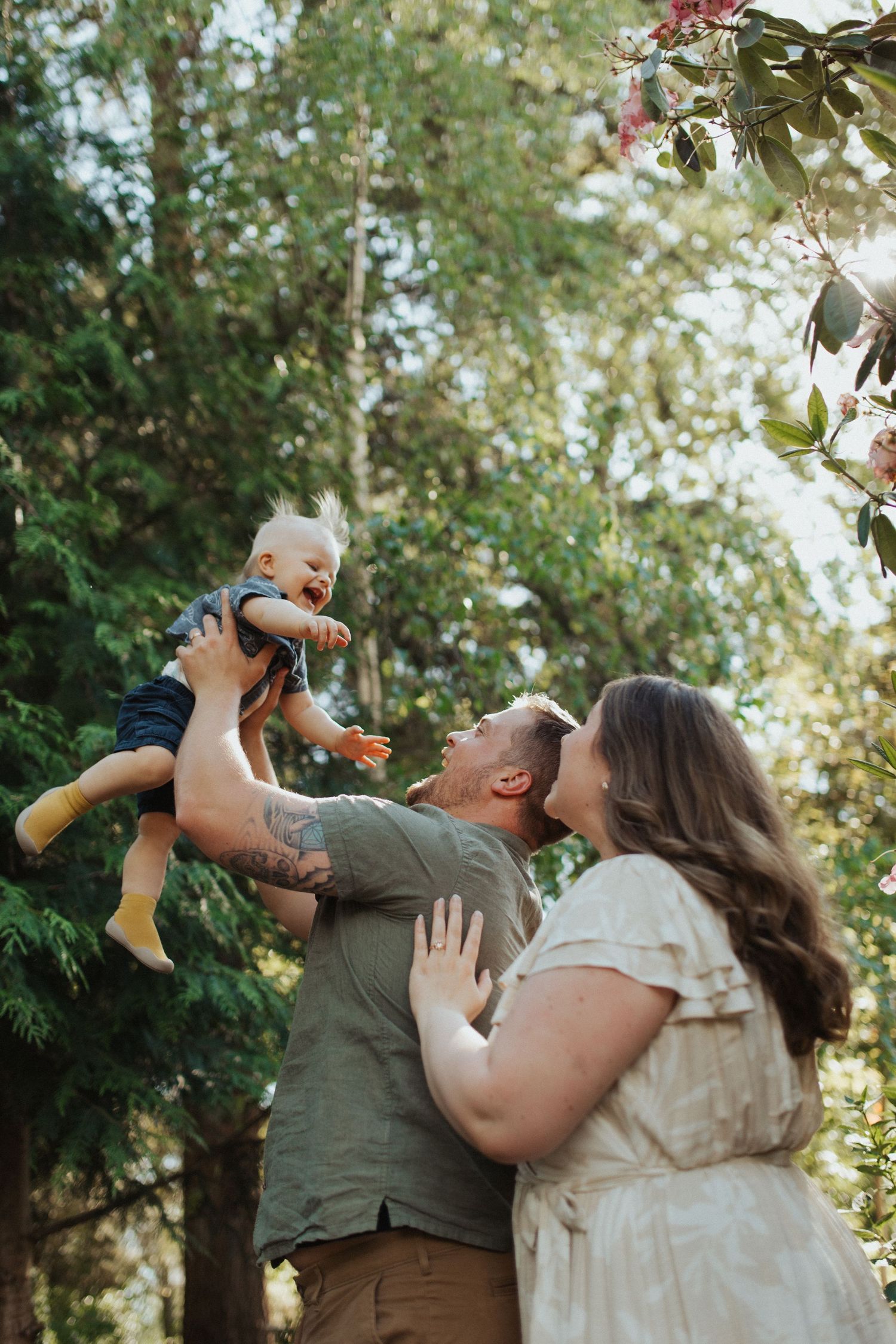 Smiling parents playfully lift their giggling baby up in a sunlit garden with blooming flowers.