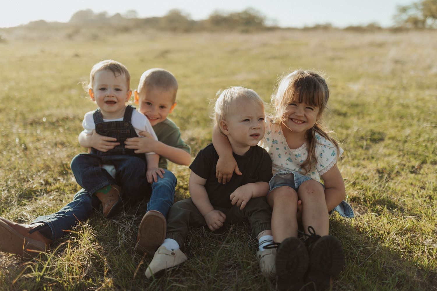 Four young children sitting together in a grassy field laughing and hugging in warm sunlight.