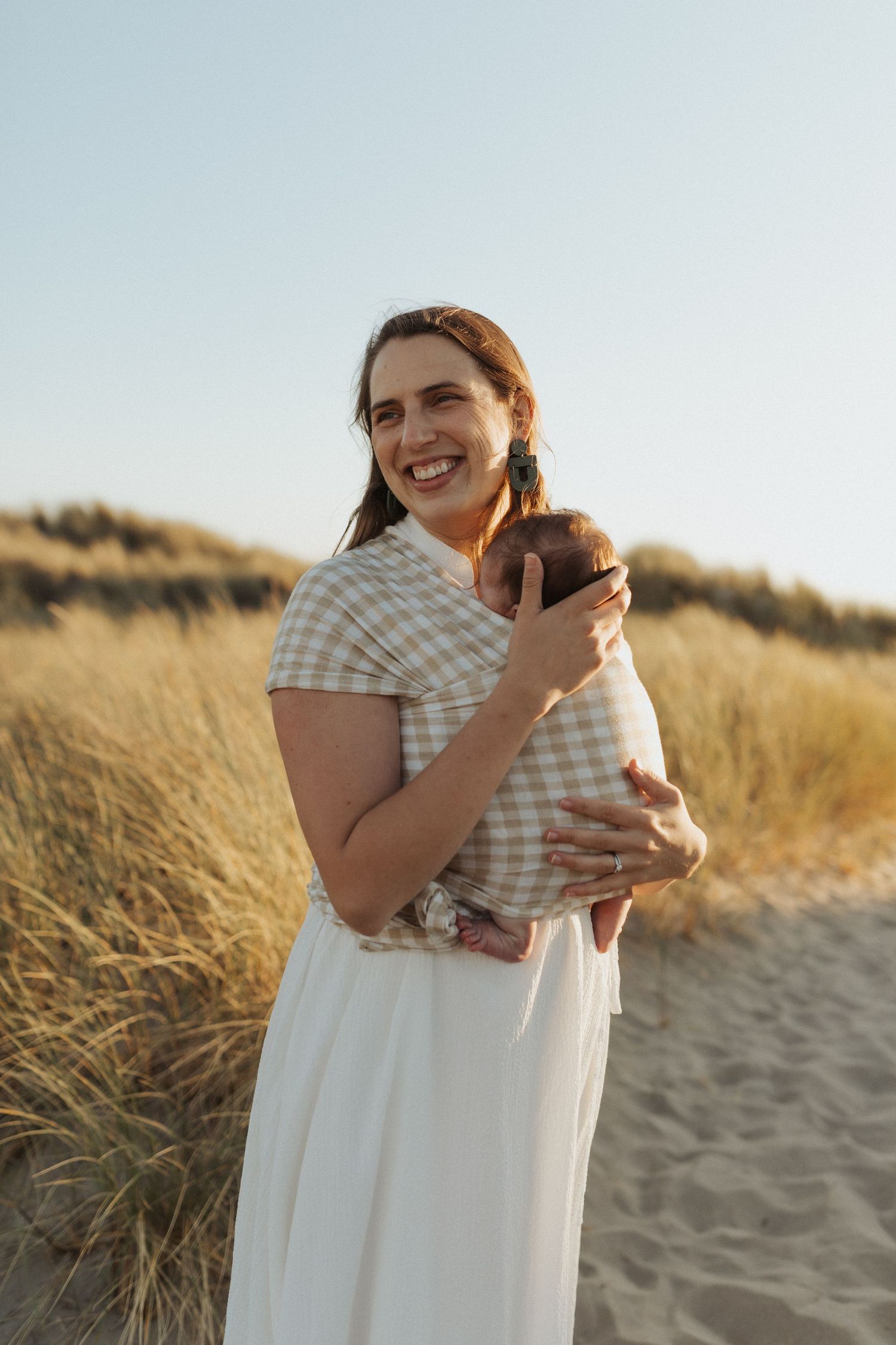 Smiling mother in white skirt and plaid top holding newborn baby on sandy beach dunes.