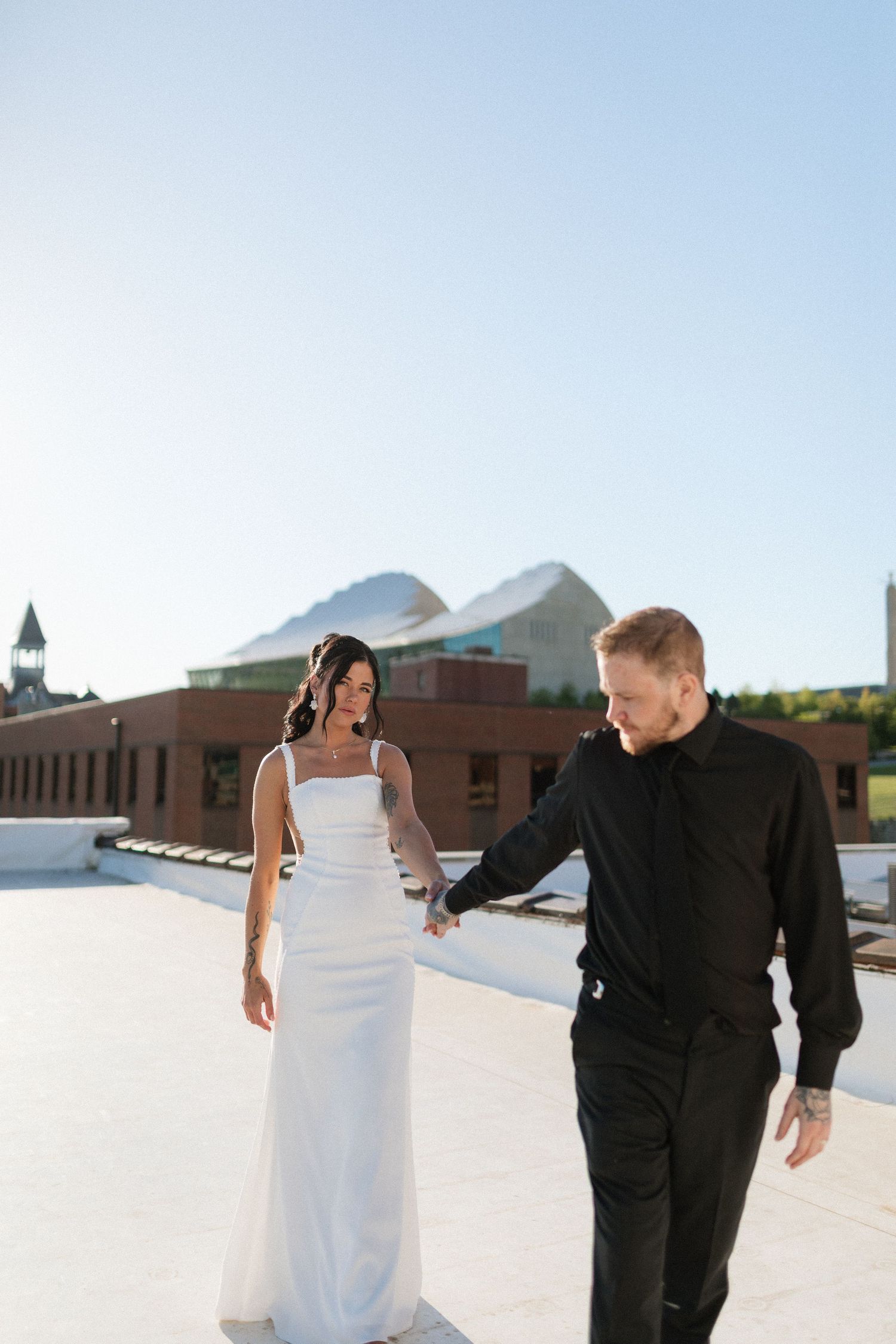 A bride in a white gown and groom in black walk hand in hand on a rooftop with a clear blue sky behind them.