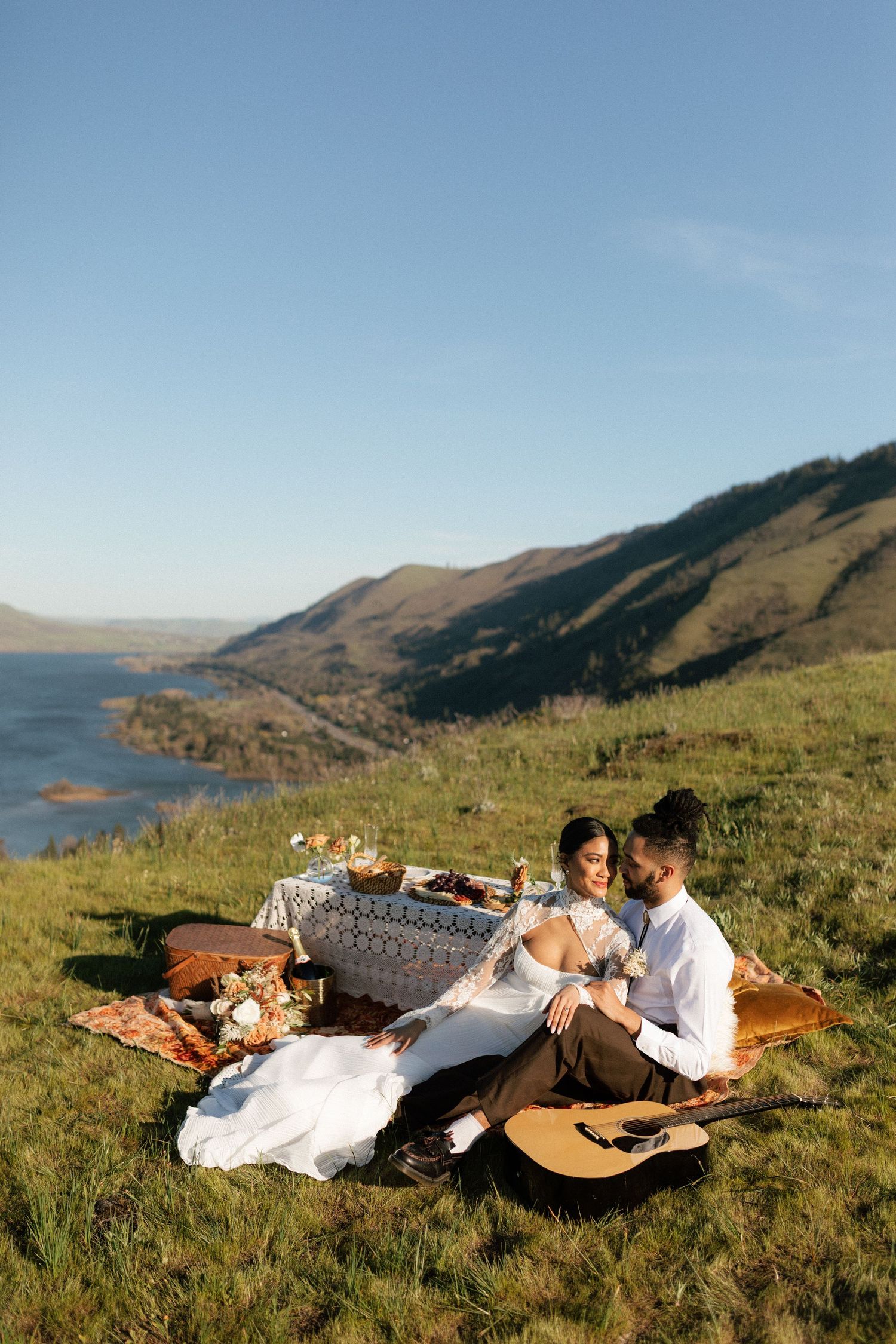 Bride and groom enjoy a boho picnic on a grassy hillside with stunning mountain lake views and a guitar nearby.