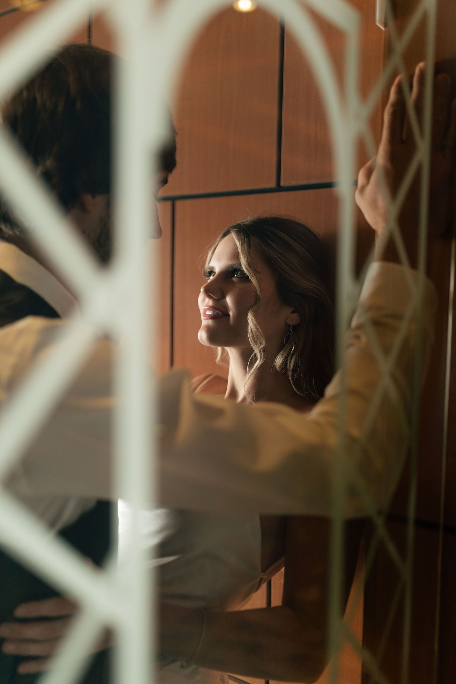 Smiling bride gazes at groom through decorative white lattice window in warm ambient light.