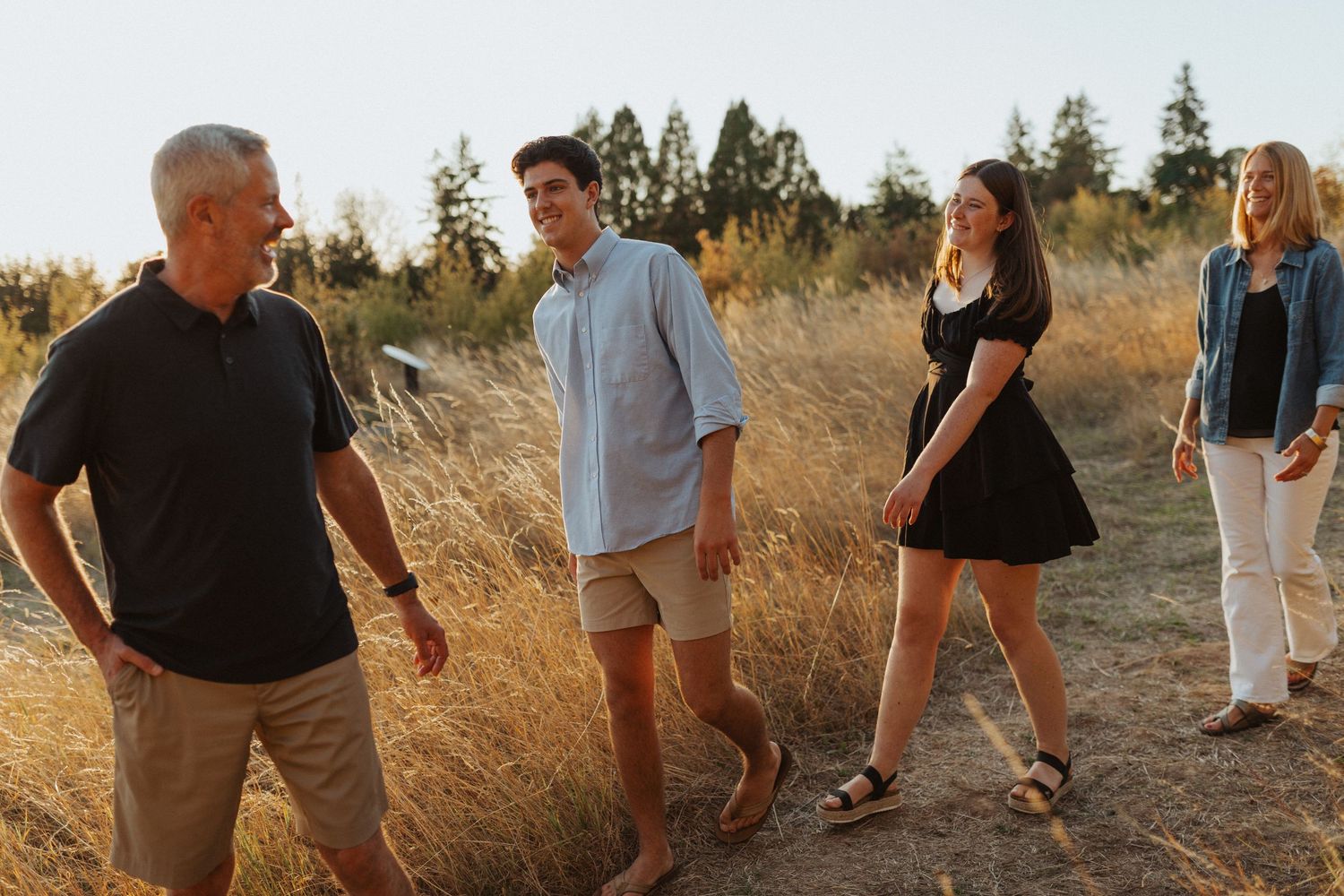 Family of four walking on a sunny dirt path through golden grass and pine trees.