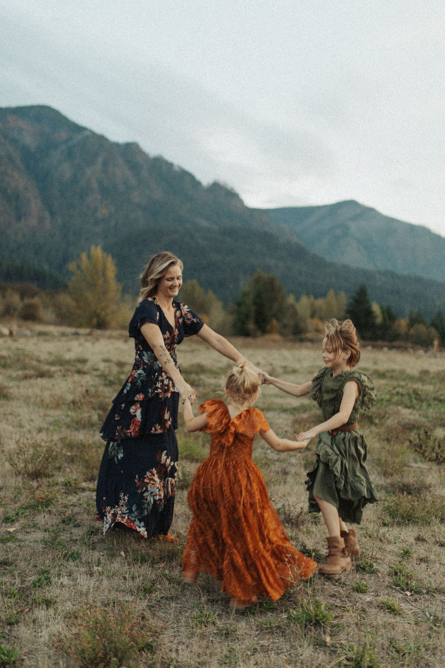 A mother and two daughters hold hands and spin together in a mountain meadow on a cloudy day.