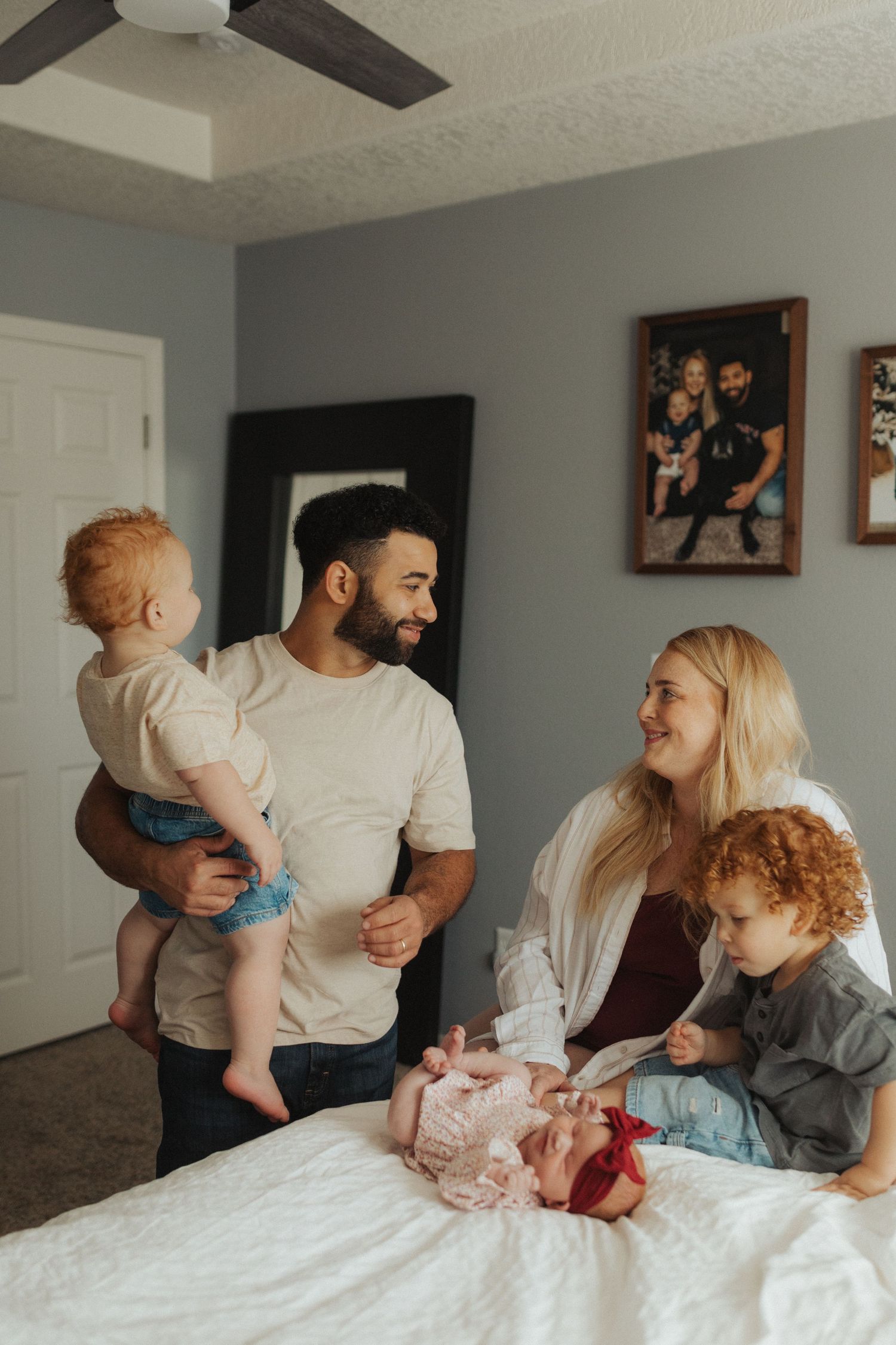A father holds a red-haired baby while a mother and toddler sit on a bed with a newborn.