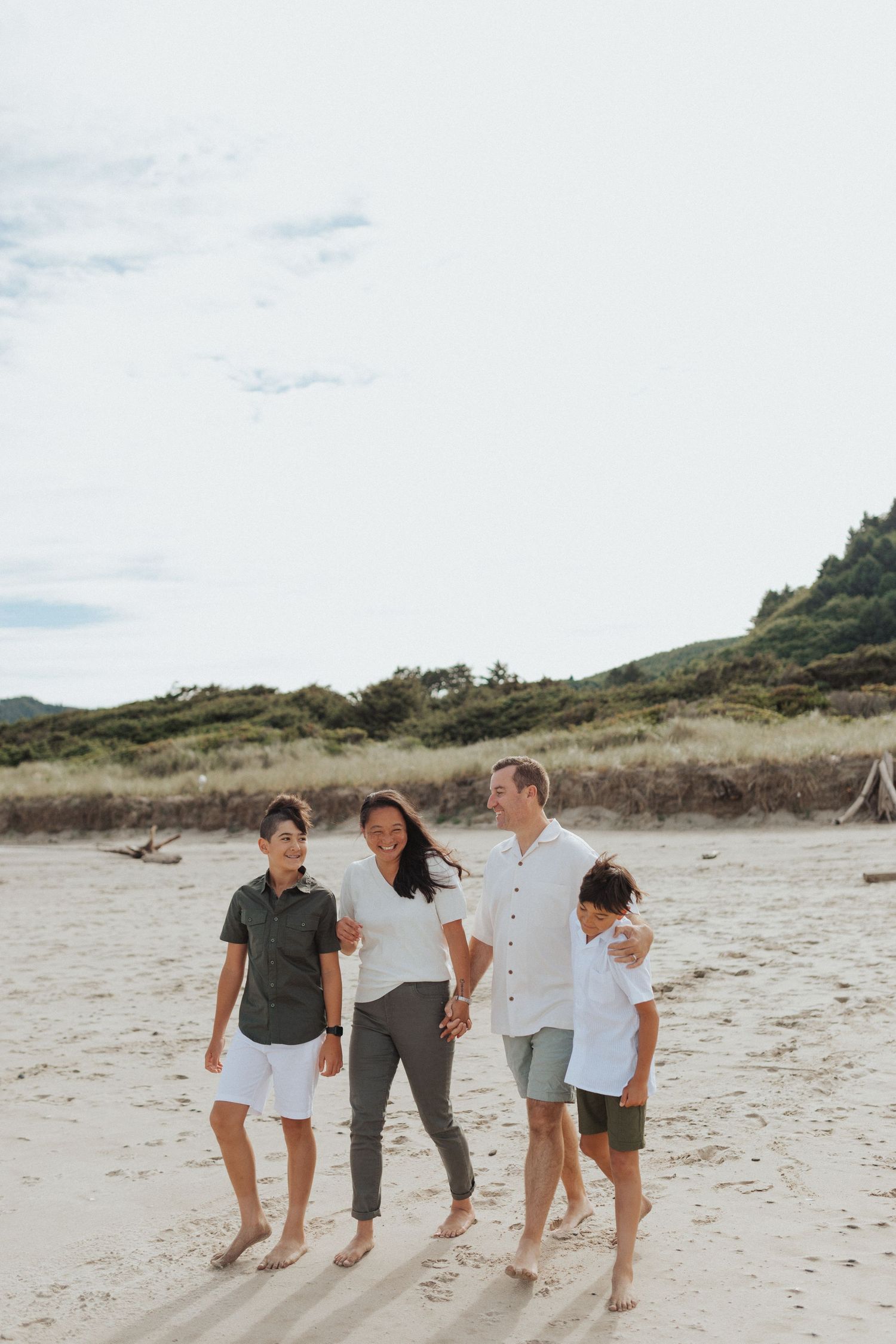 Family of four walking barefoot together on a sandy beach, smiling and enjoying the outdoors.