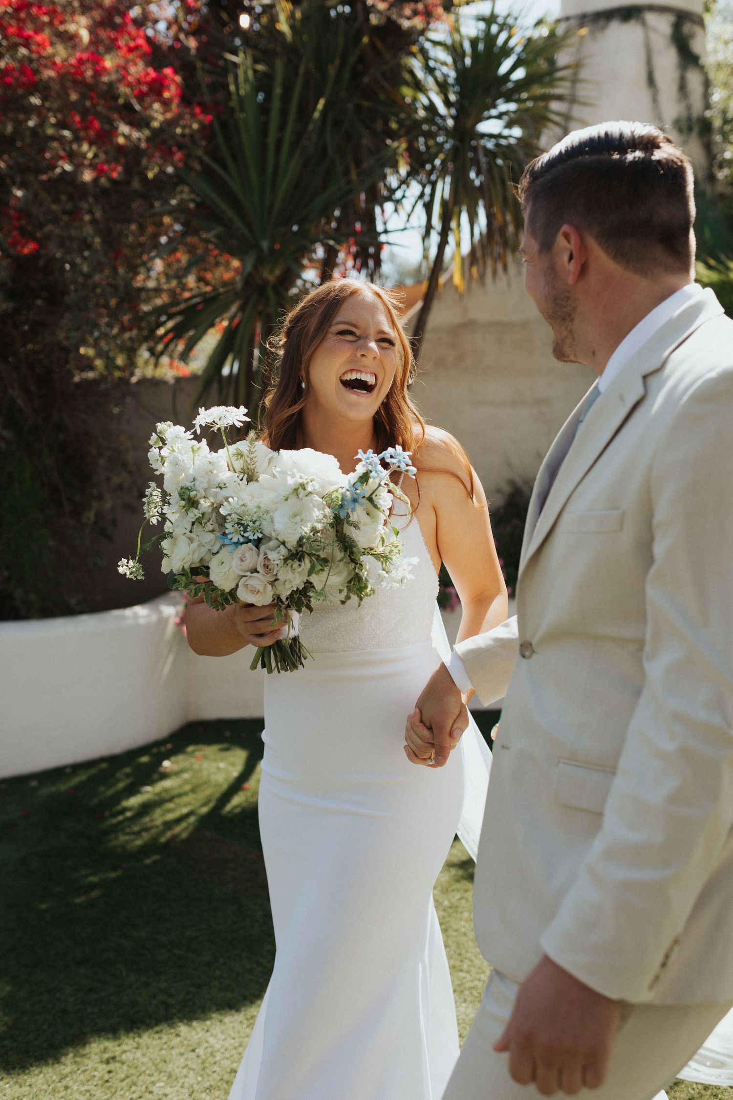 Laughing bride in a white gown holds a blue and white bouquet while holding hands with groom in cream suit outdoors.
