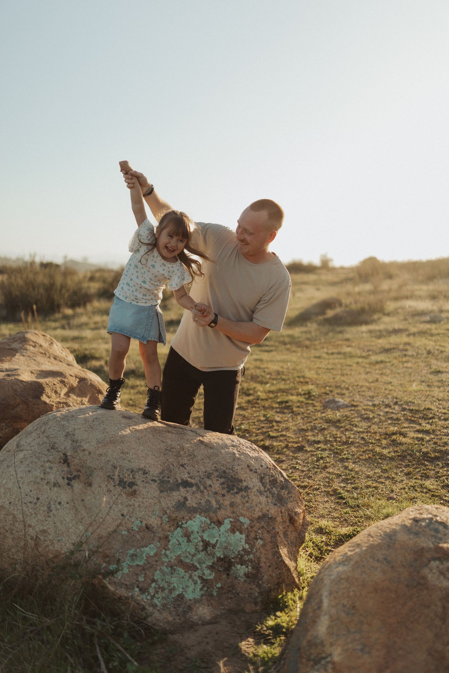 Father helping young daughter balance on large rock in sunny open field during outdoor family session.