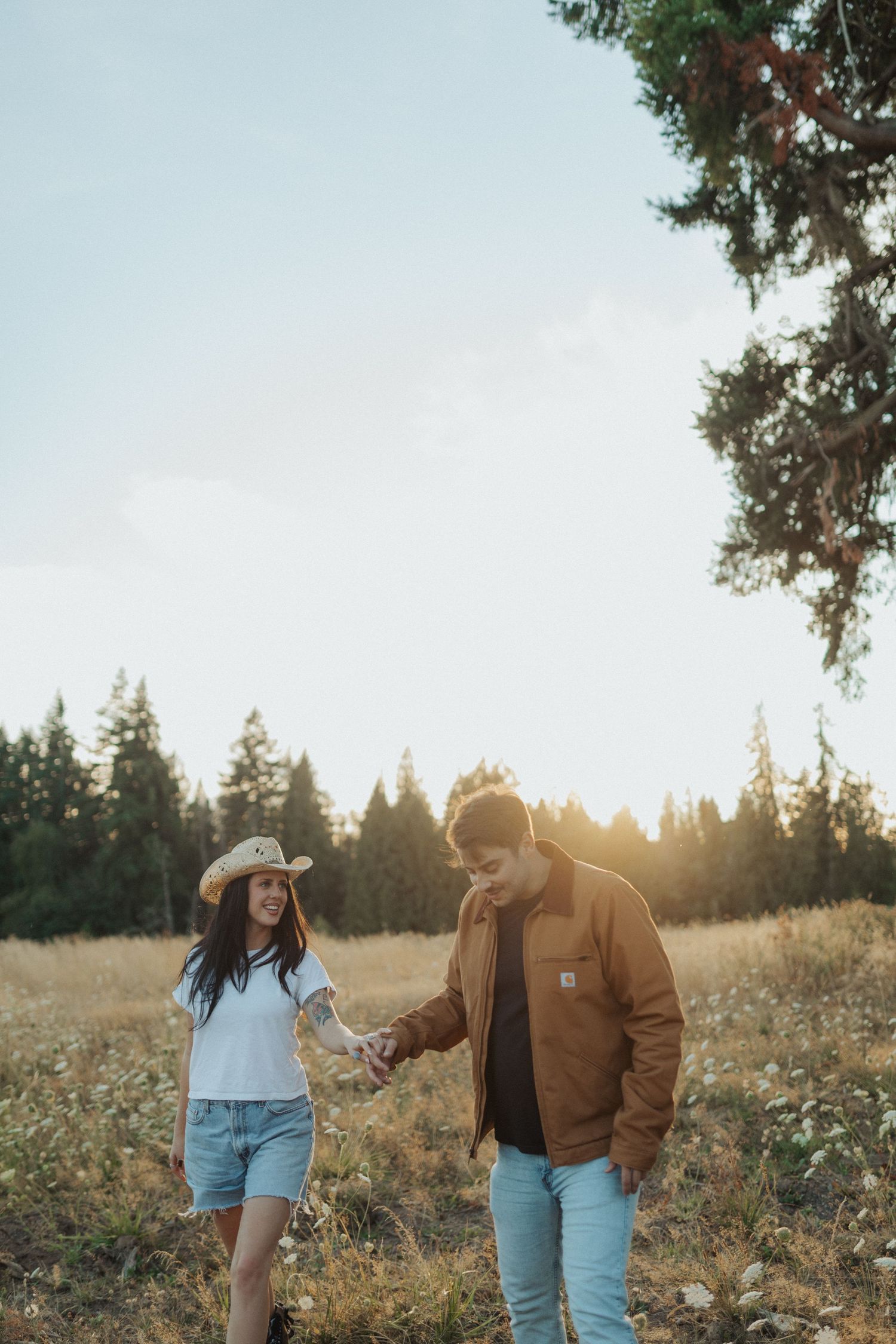 A couple holds hands walking through a sunlit field with tall pine trees glowing in the background.