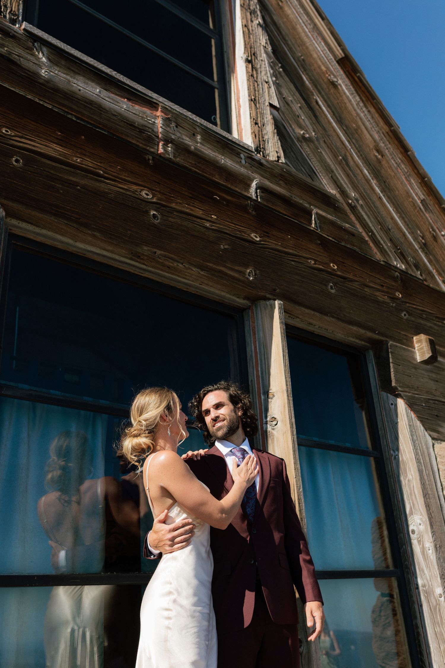 Bride and groom embrace outside rustic wooden building on sunny wedding day.