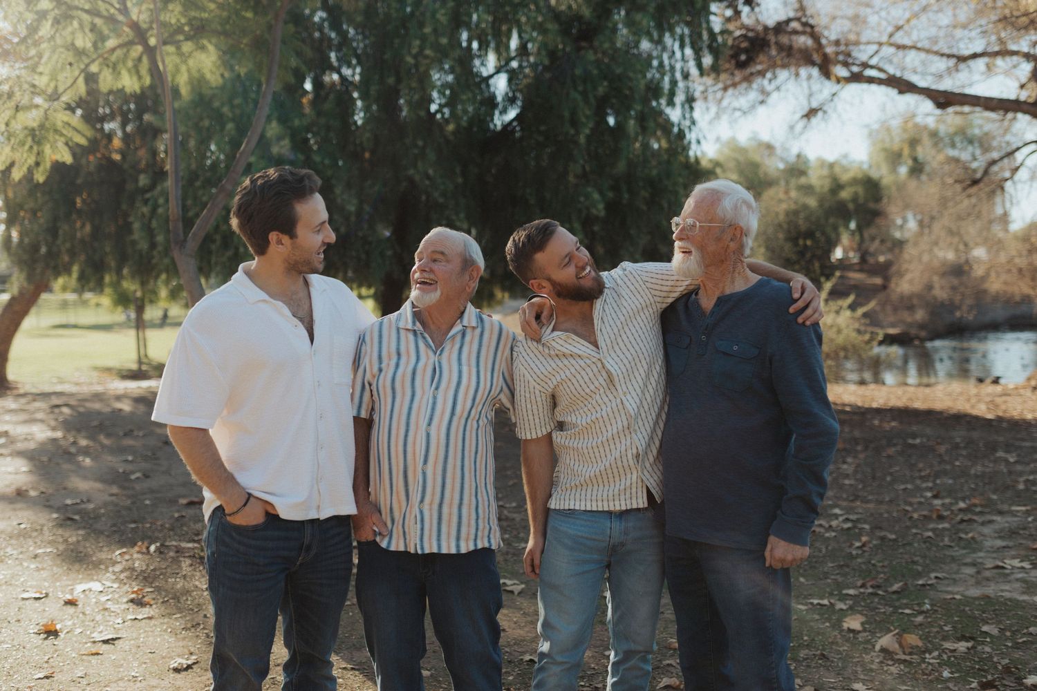 Four men of different ages laugh and walk together arm in arm in a sunny park.