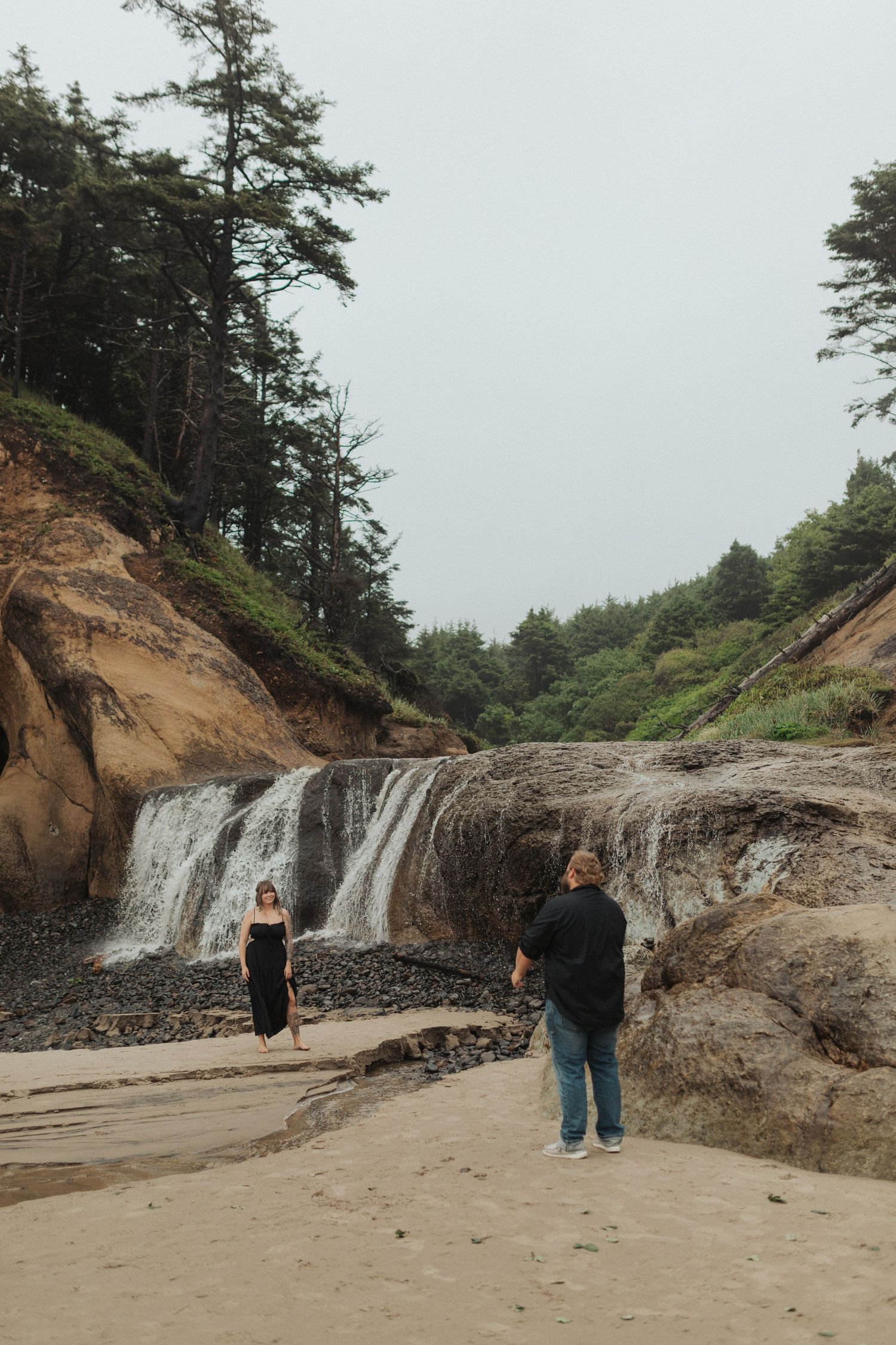 Photographer capturing a woman in black dress standing near a waterfall on a sandy beach.