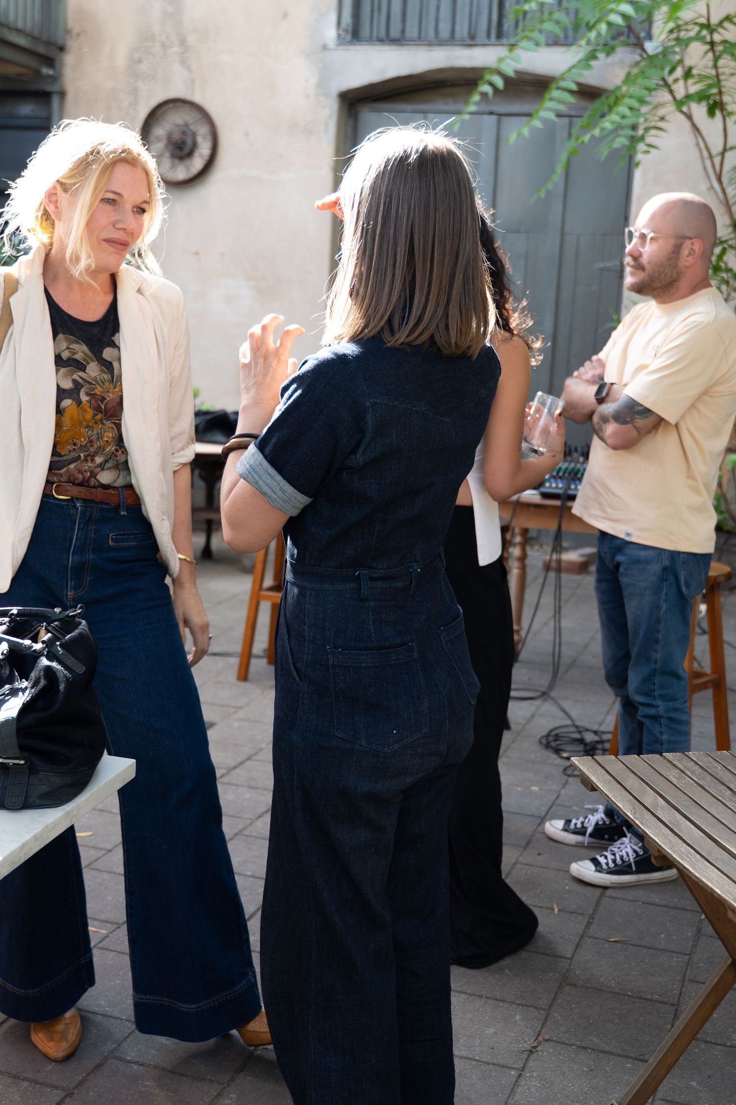 People socializing outdoors at a casual gathering, with a woman in denim overalls gesturing while talking to a blonde woman.