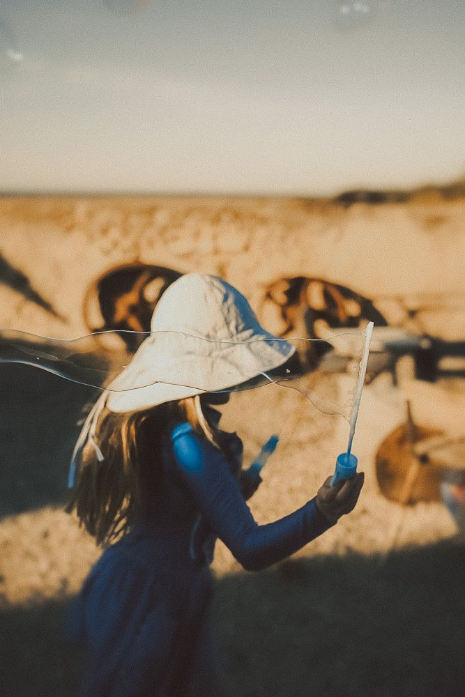 A girl in a white sun hat and blue dress holds a stick while standing in a sandy desert landscape at golden hour.