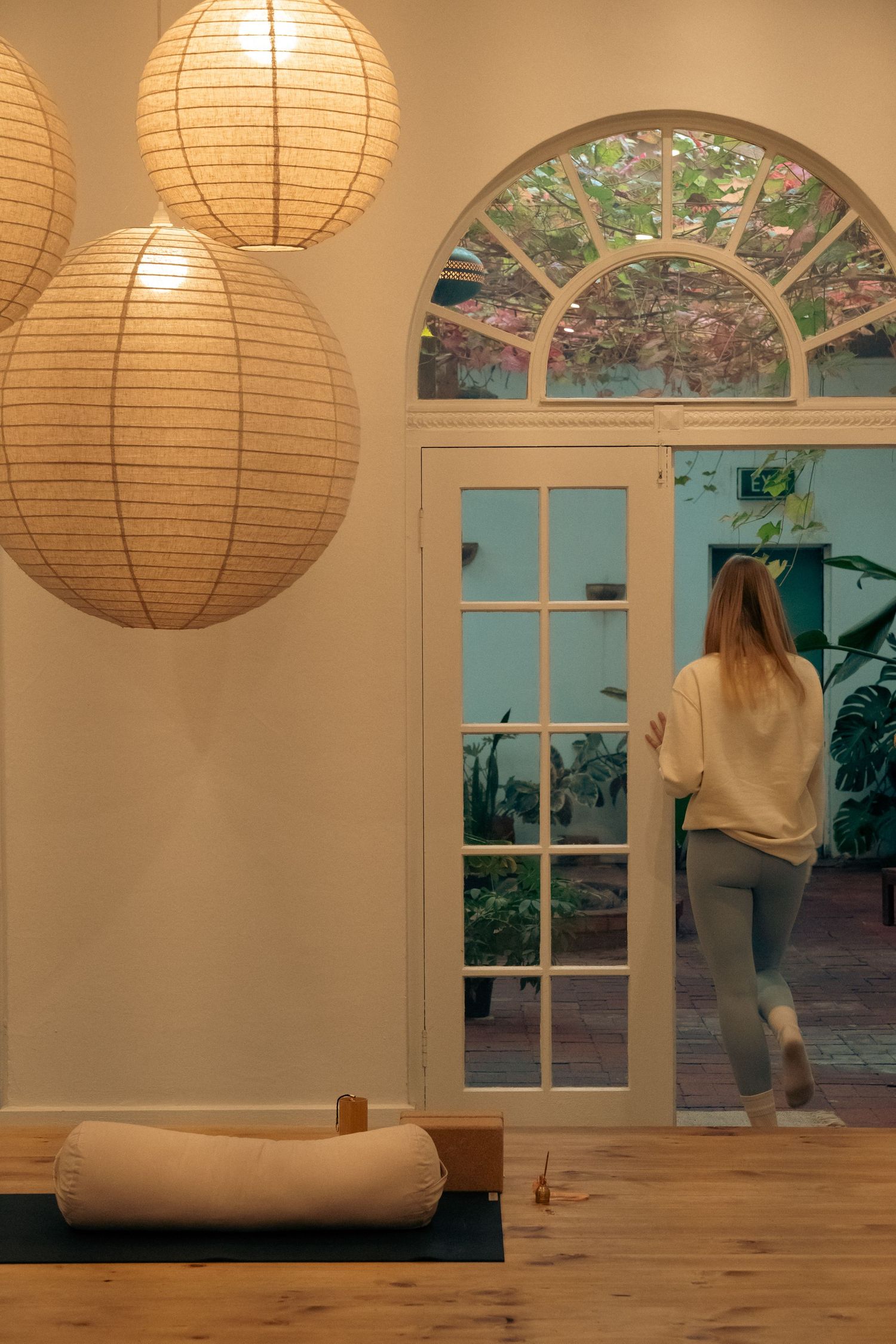 Woman walking toward arched French doors in a serene yoga studio with paper lantern lights and a meditation bolster on floor.