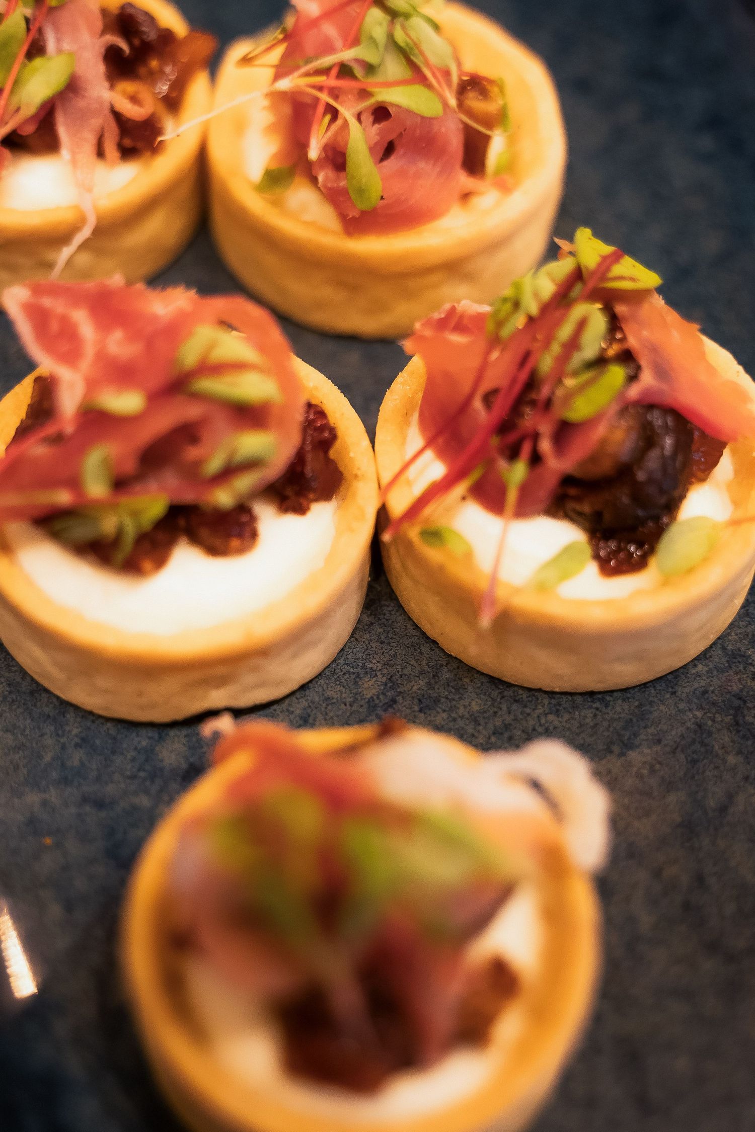Elegant canape tartlets topped with cured meat, cream cheese, chutney, and microgreens on a slate board.