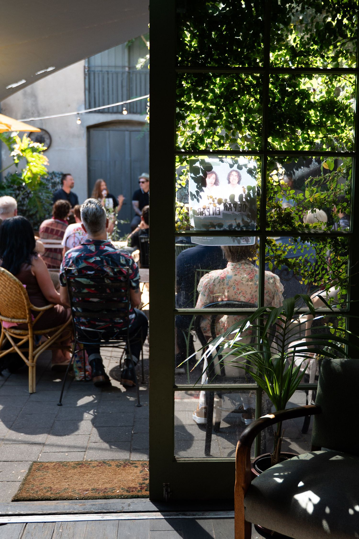 People dining in a sunlit outdoor patio garden cafe, viewed through a green-framed glass door with lush foliage.