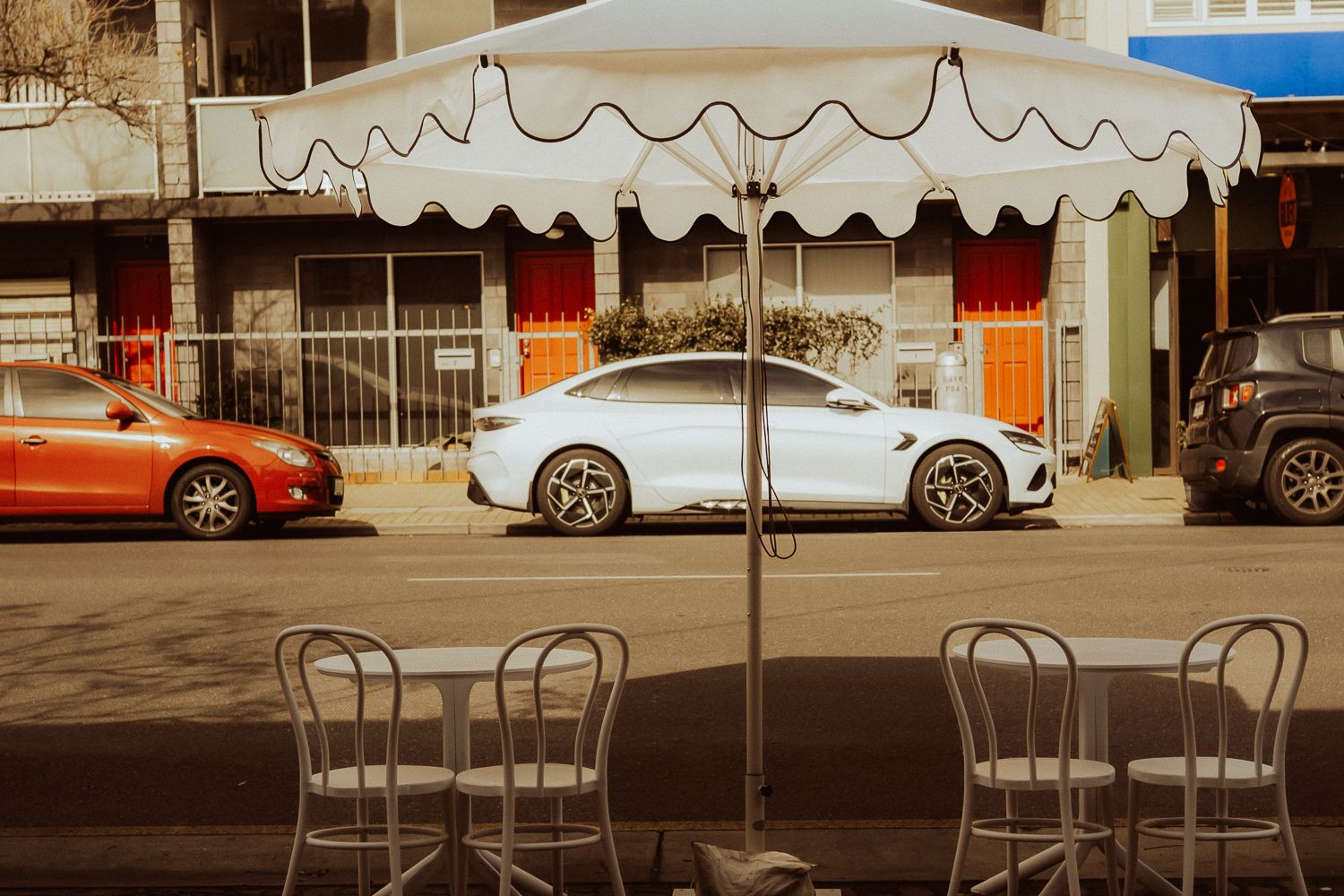 White luxury car parked on a city street viewed from a sidewalk cafe with umbrella and chairs.