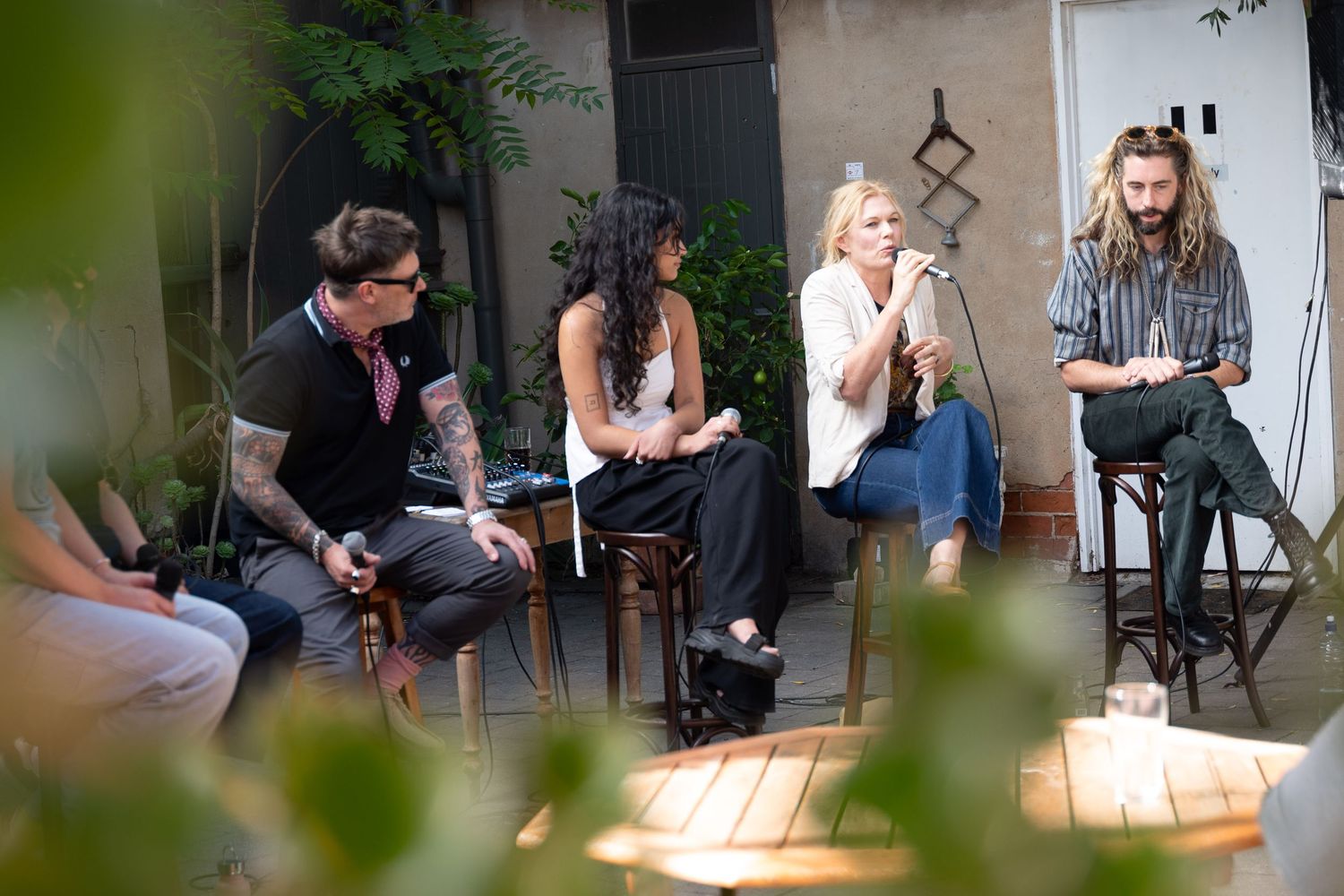 Four people engaged in a panel discussion outdoors, seated on stools in a courtyard surrounded by lush green plants.