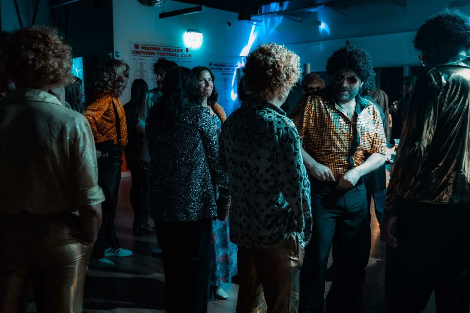 Group of people in 1970s retro outfits socializing at a dimly lit disco party venue.
