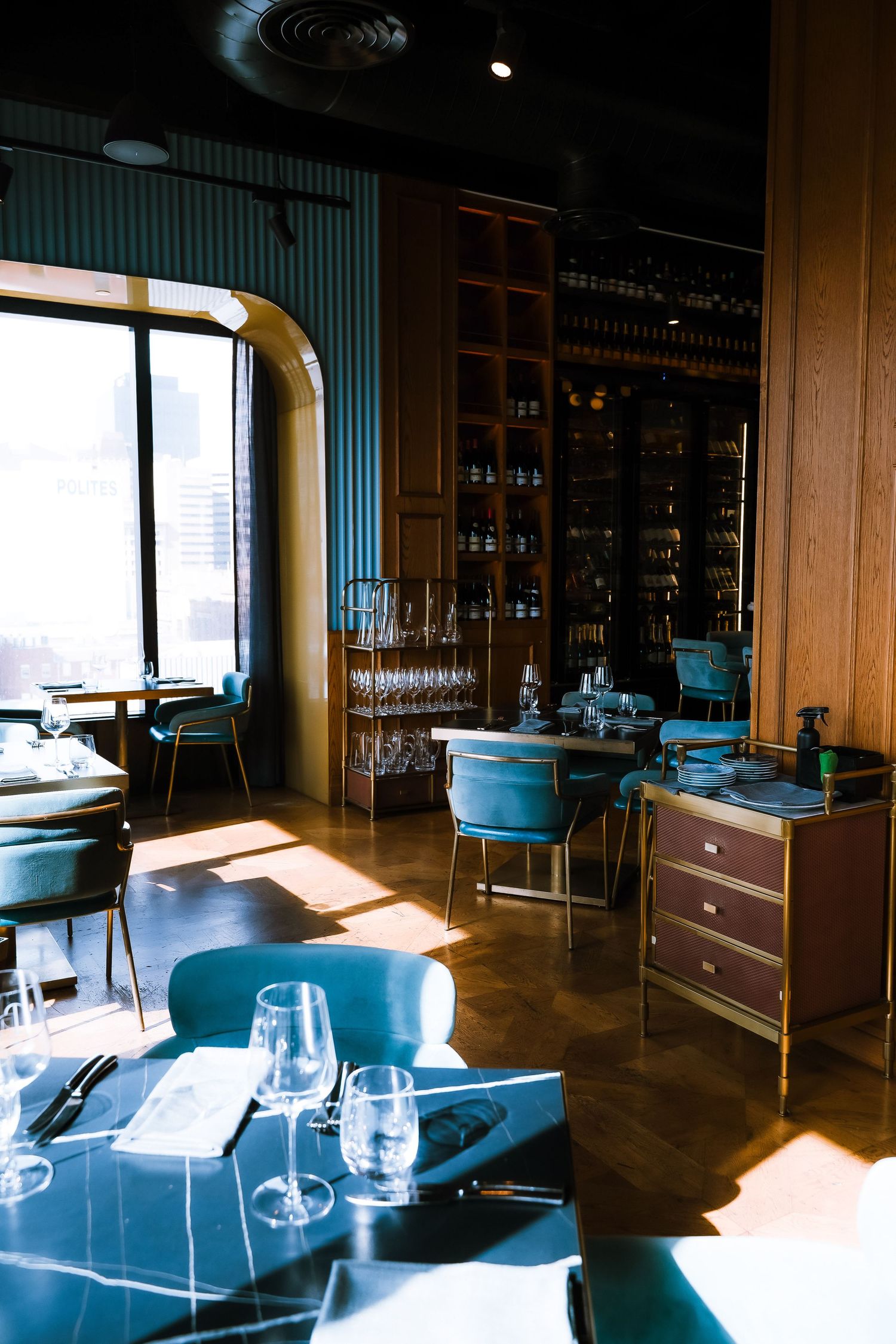Elegant restaurant interior with blue velvet chairs, set tables with wine glasses, wooden cabinets, and natural light from arched windows.