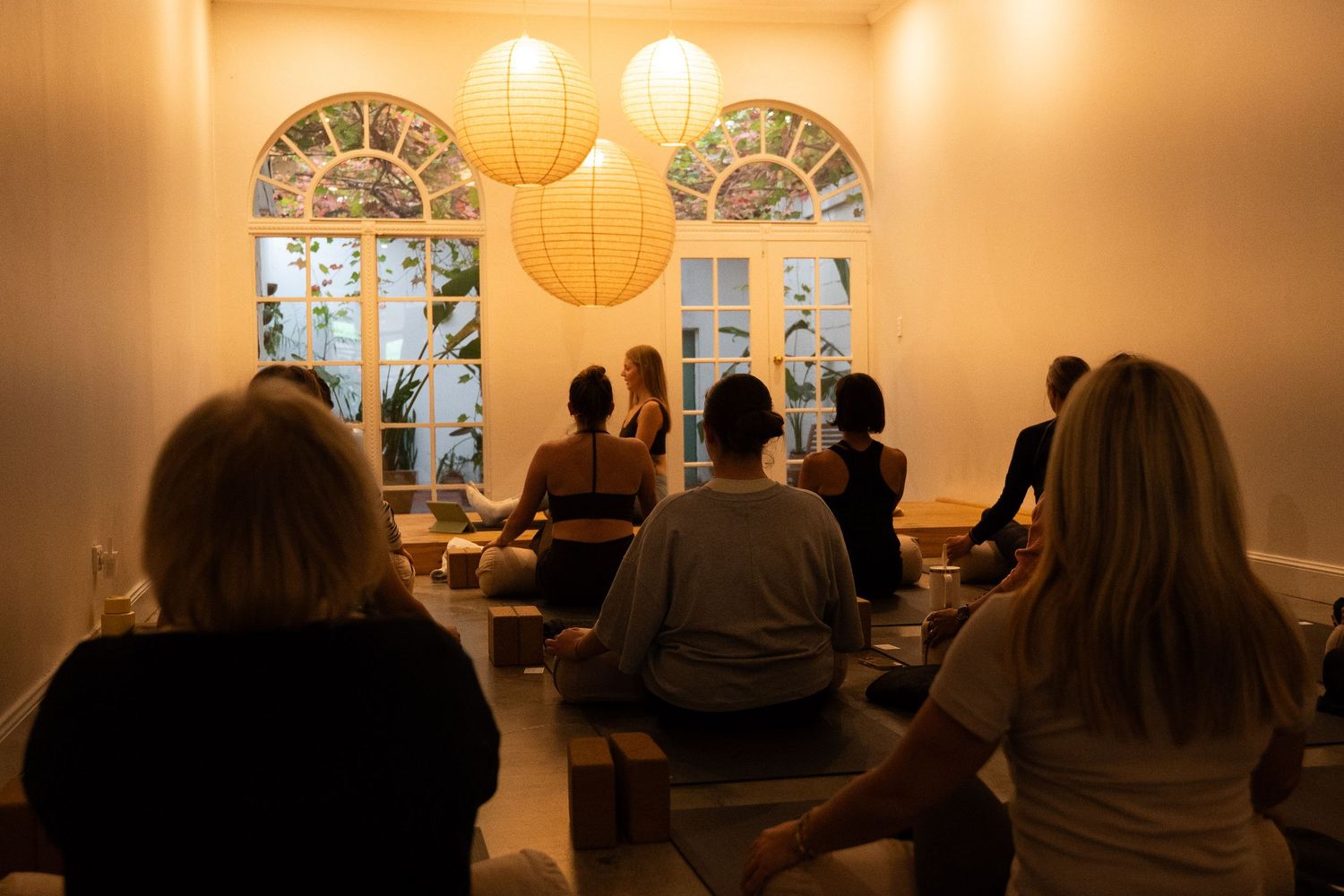 Group meditation session in a warmly lit room with paper lantern lights and arched stained glass windows.