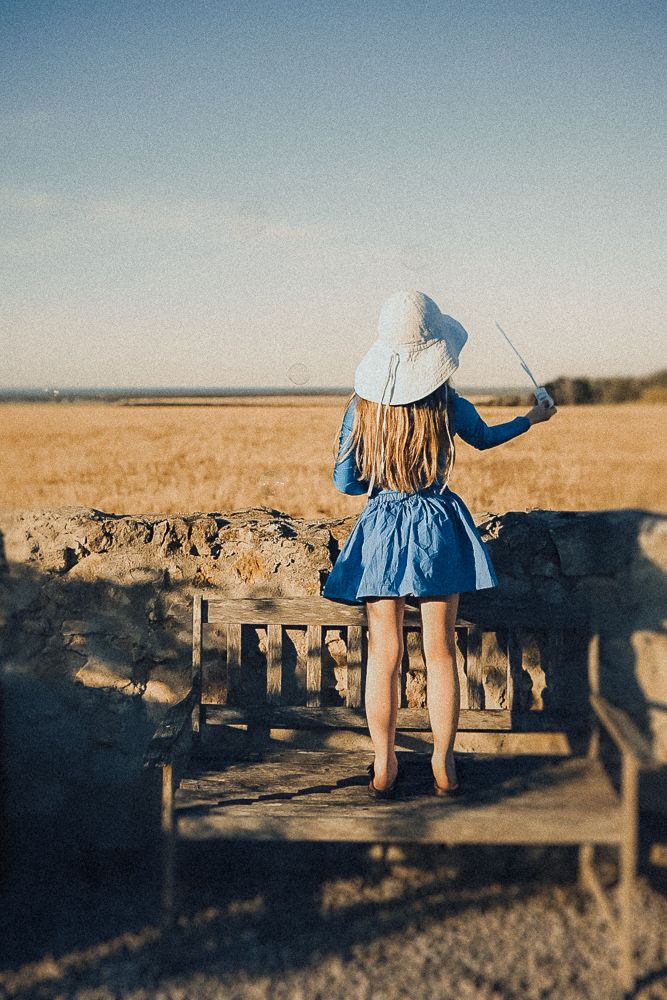 Young woman in blue dress and sun hat standing on wooden bench overlooking golden field landscape.