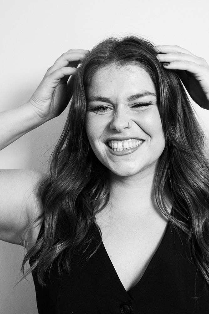 Black and white portrait of a laughing young woman with long wavy hair, hands in hair, wearing a black top.