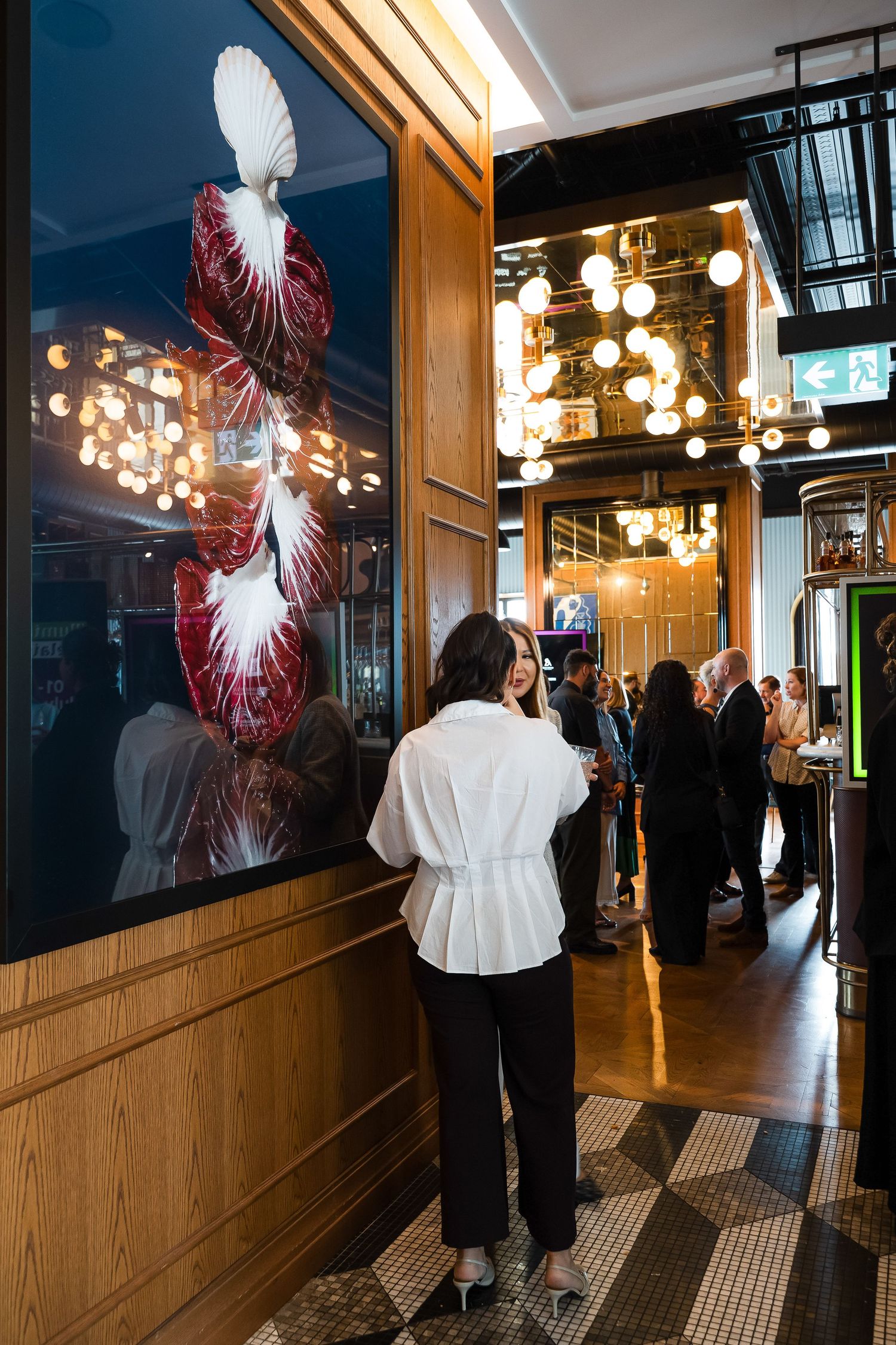 Woman in white jacket viewing floral art display in elegant restaurant bar with globe pendant lights.