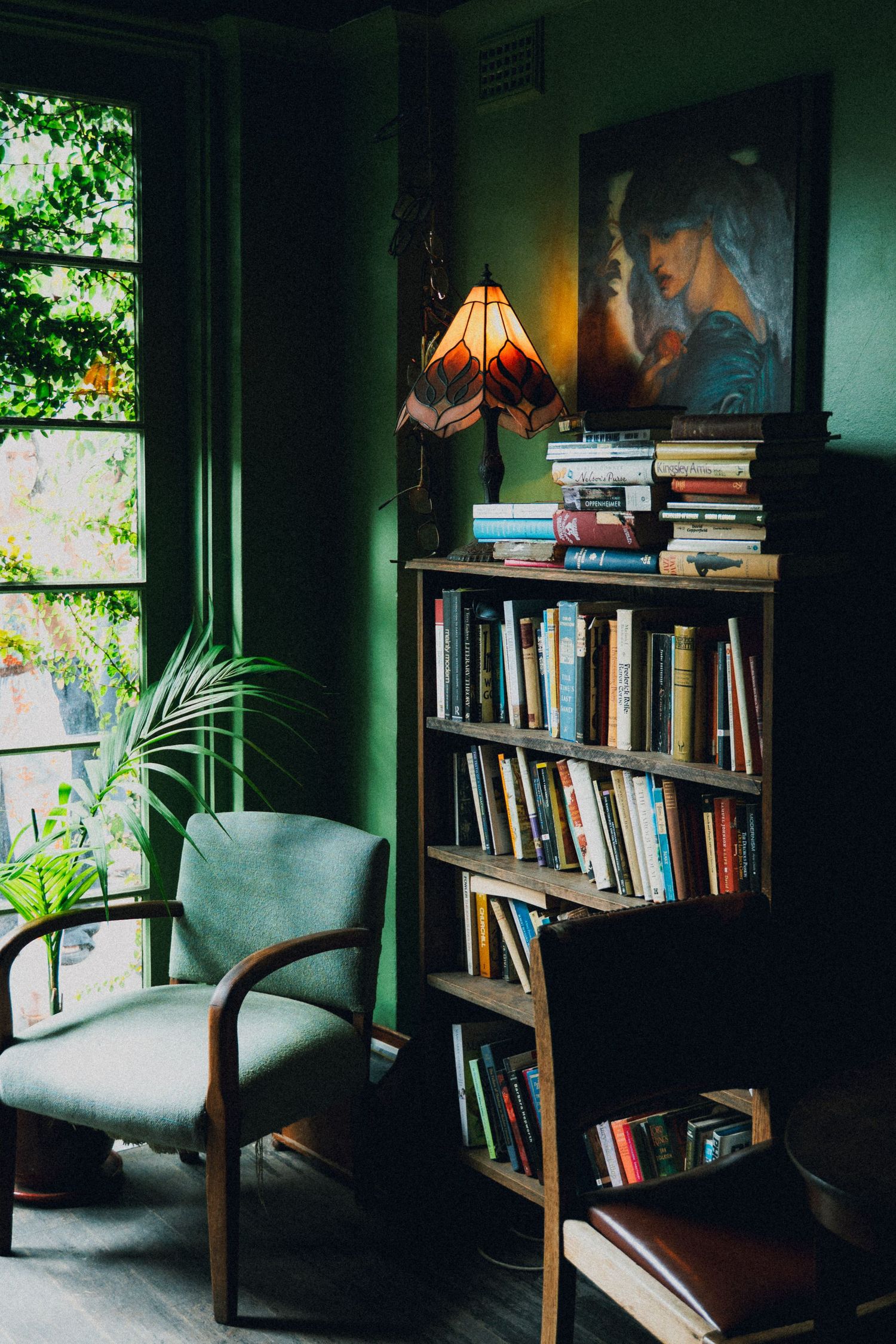 Cozy reading nook with green walls, wooden bookshelf filled with books, a lit stained glass lamp, and a vintage armchair.