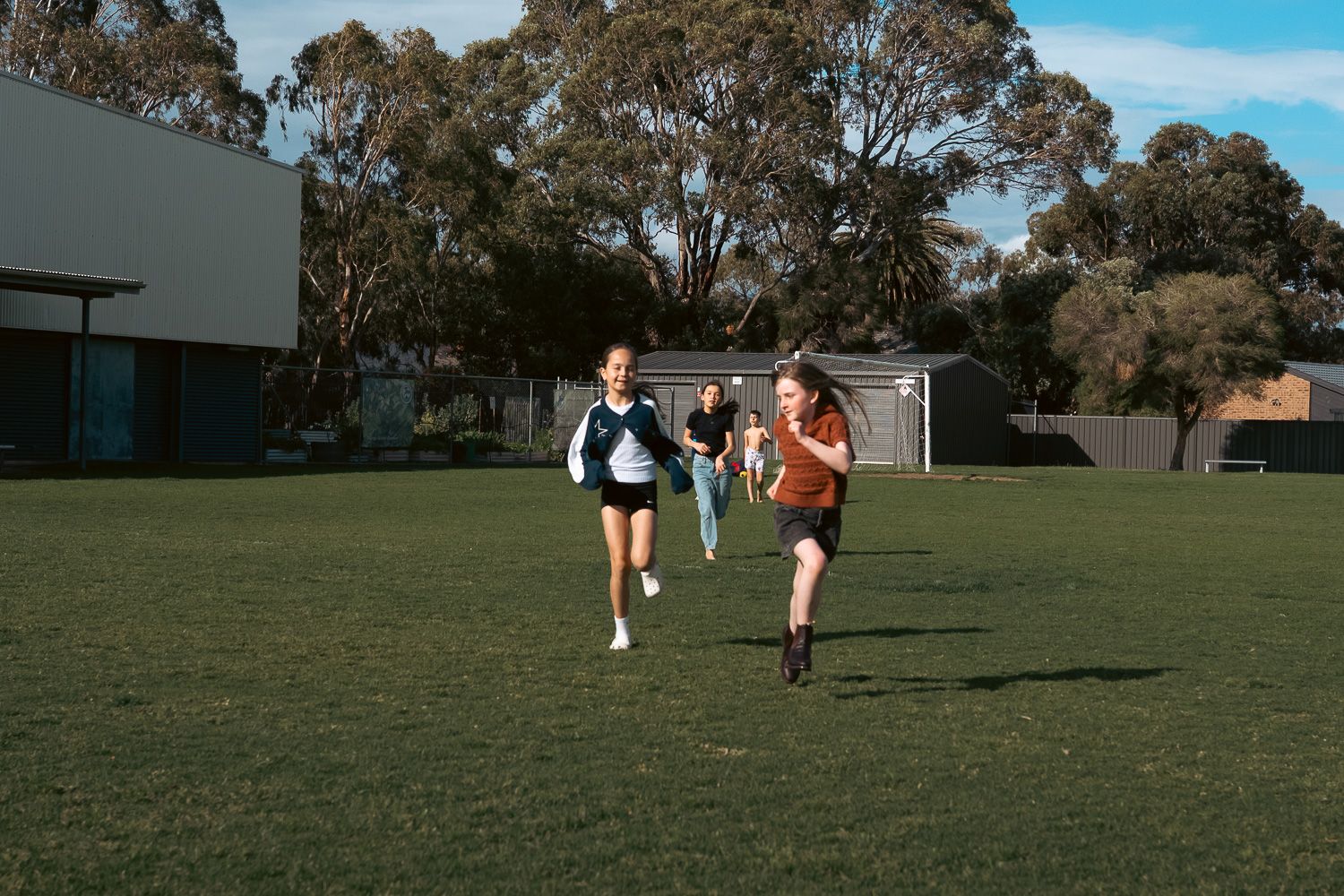 Girls playing football on a green grass field at school with trees in the background on a sunny day.