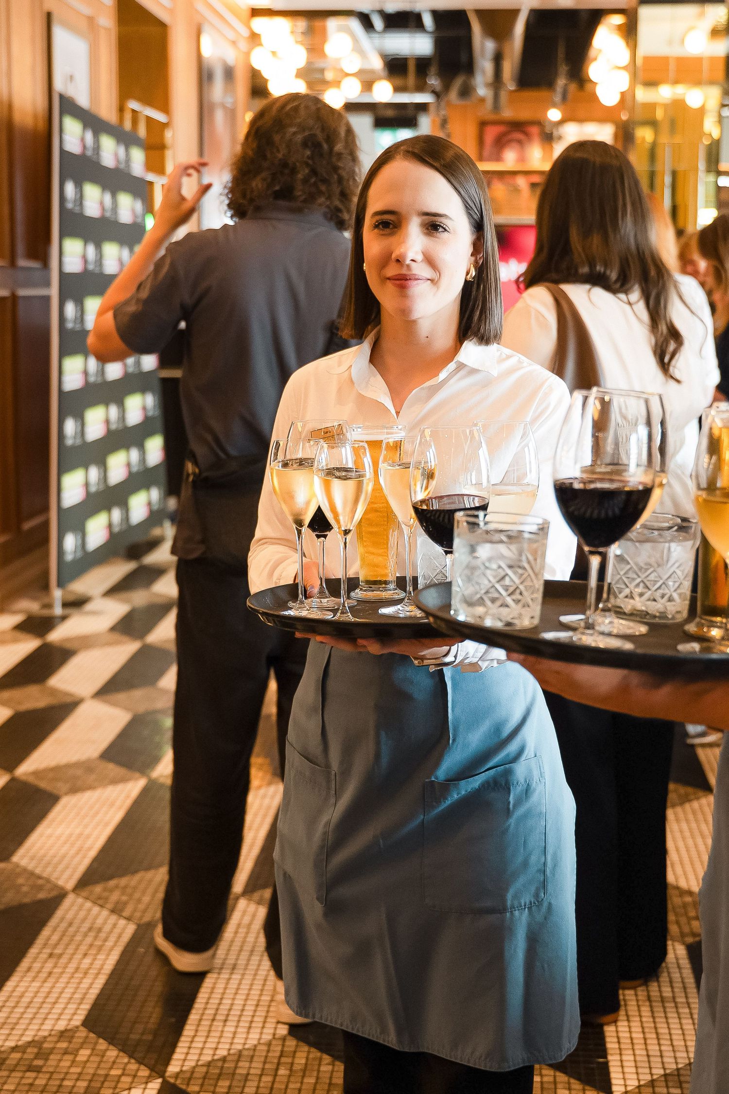 A smiling waitress in a white shirt and blue apron carries a tray of wine glasses in an elegant restaurant.