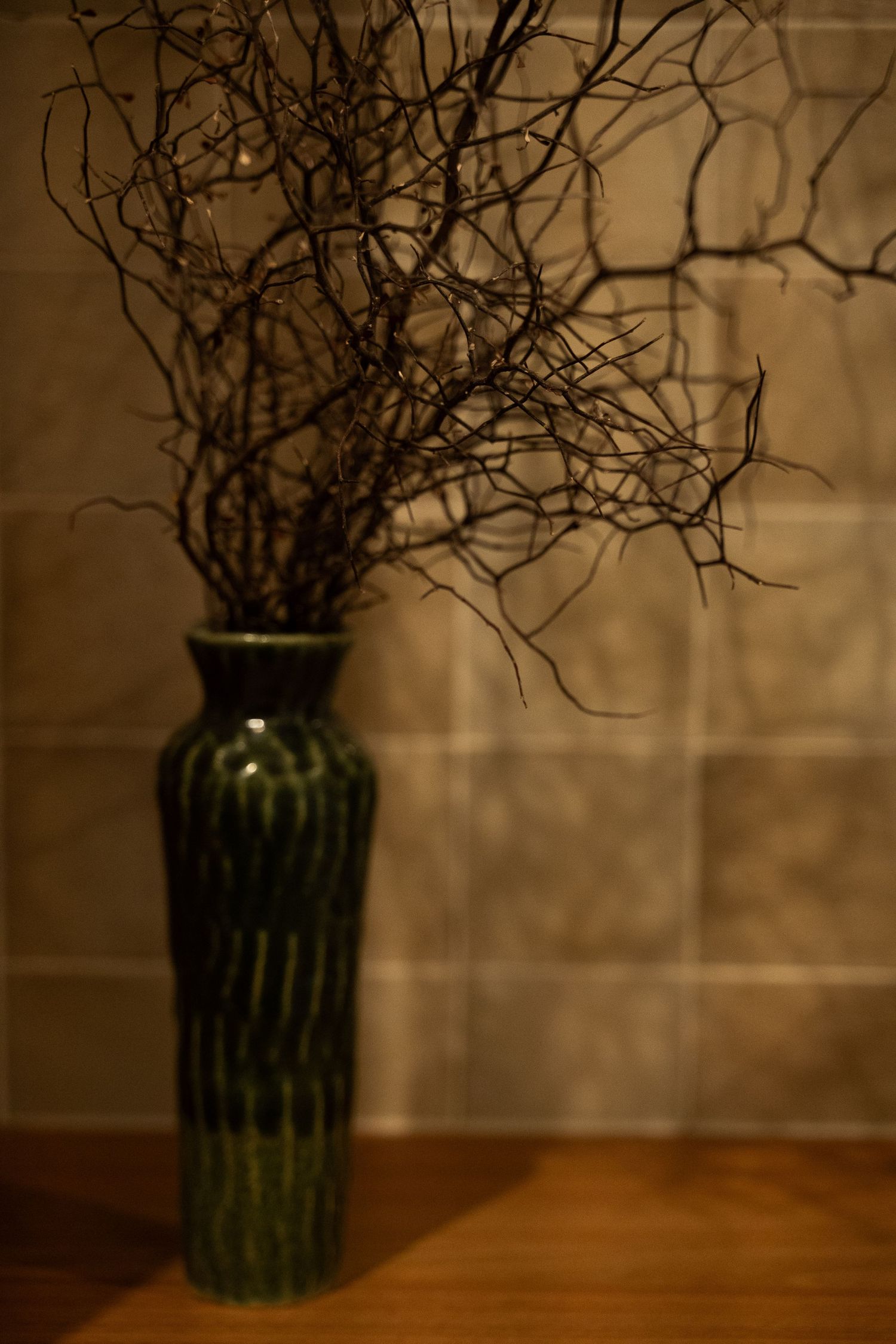 A tall green ceramic vase with twisted bare branches displayed on a wooden surface against a tiled wall.
