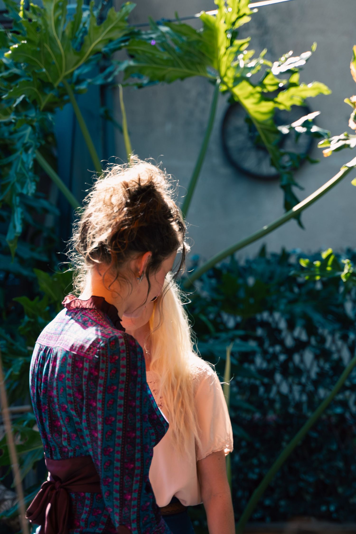 Woman with blonde hair in bun wearing floral dress stands in sunlit garden surrounded by lush green plants.