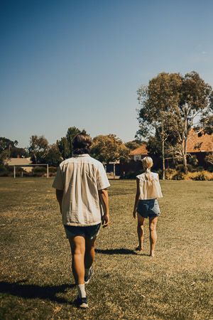 Two people walking across a sunny green field toward trees and houses in the distance.