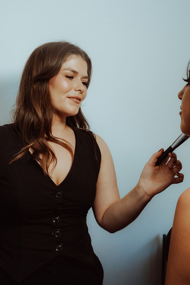 A makeup artist in a black dress applies lipstick to a client in a bright studio setting.