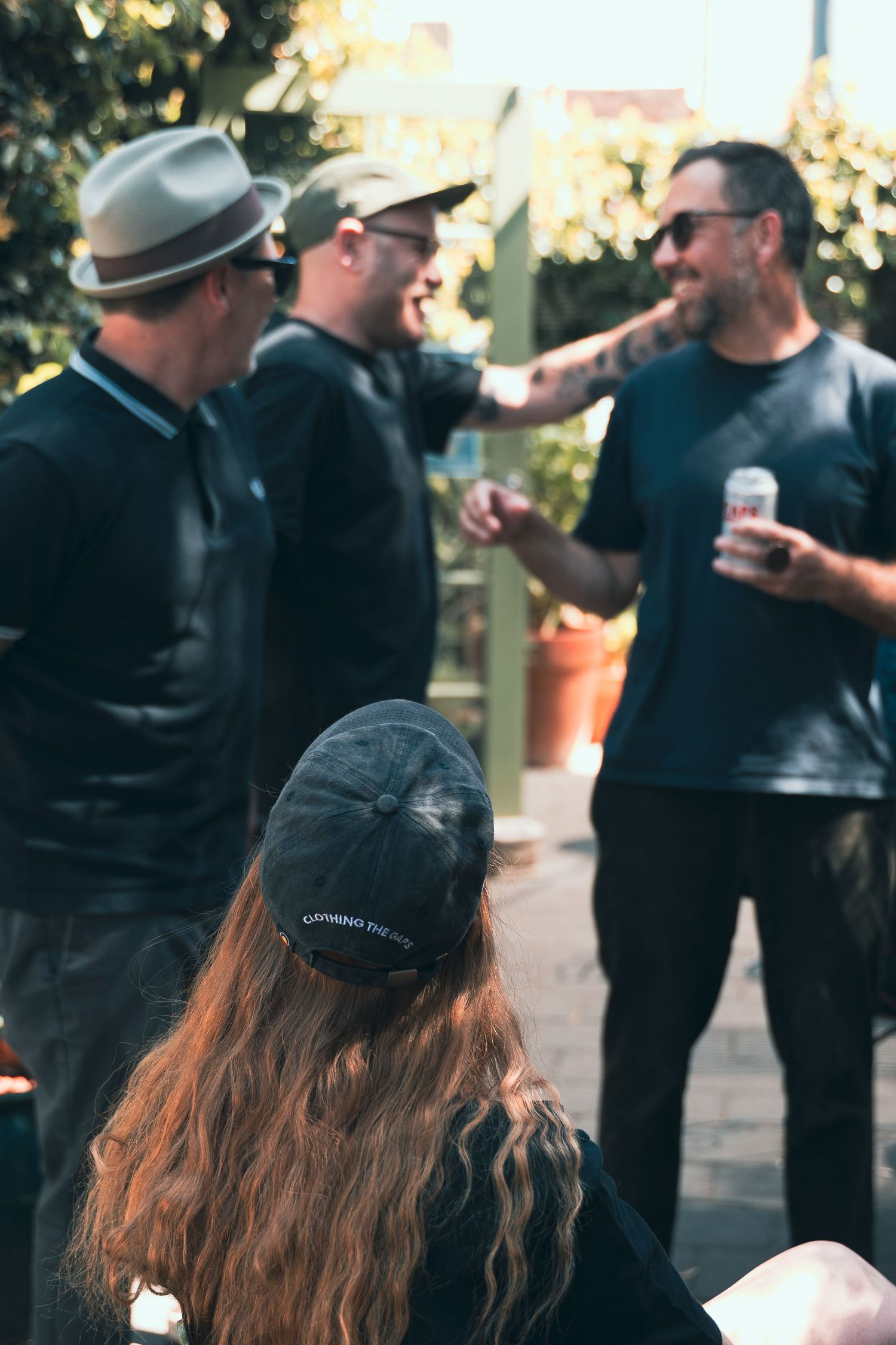 Group of friends socializing outdoors, woman with long red hair wearing black cap in foreground, men laughing behind her.