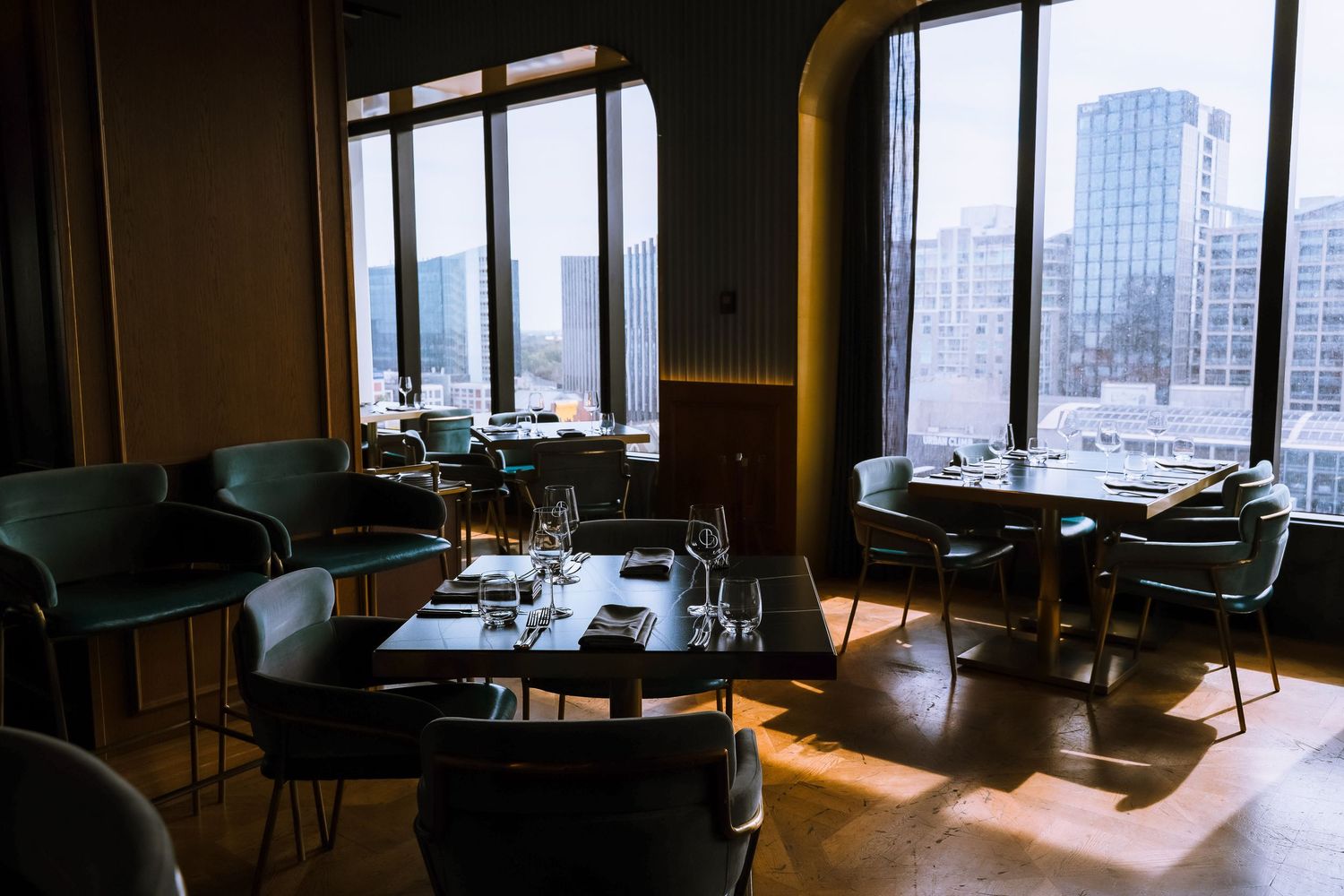 Elegant restaurant dining room with green velvet chairs, dark tables, and floor-to-ceiling windows overlooking a city skyline.