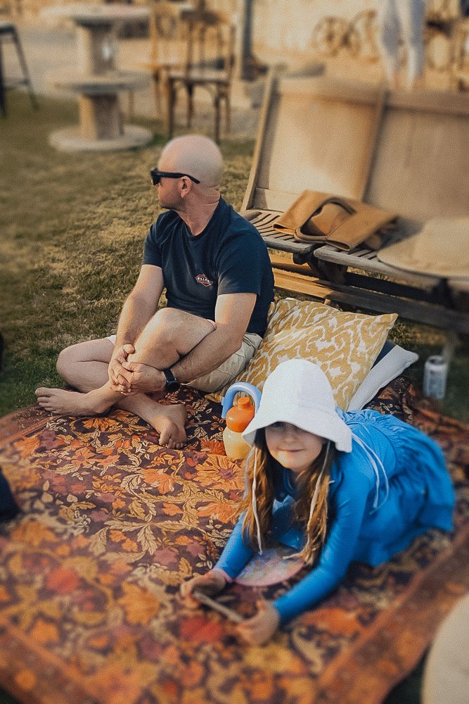 A bald man in sunglasses sits cross-legged on a patterned rug outdoors while a young girl in a white hat reads nearby.