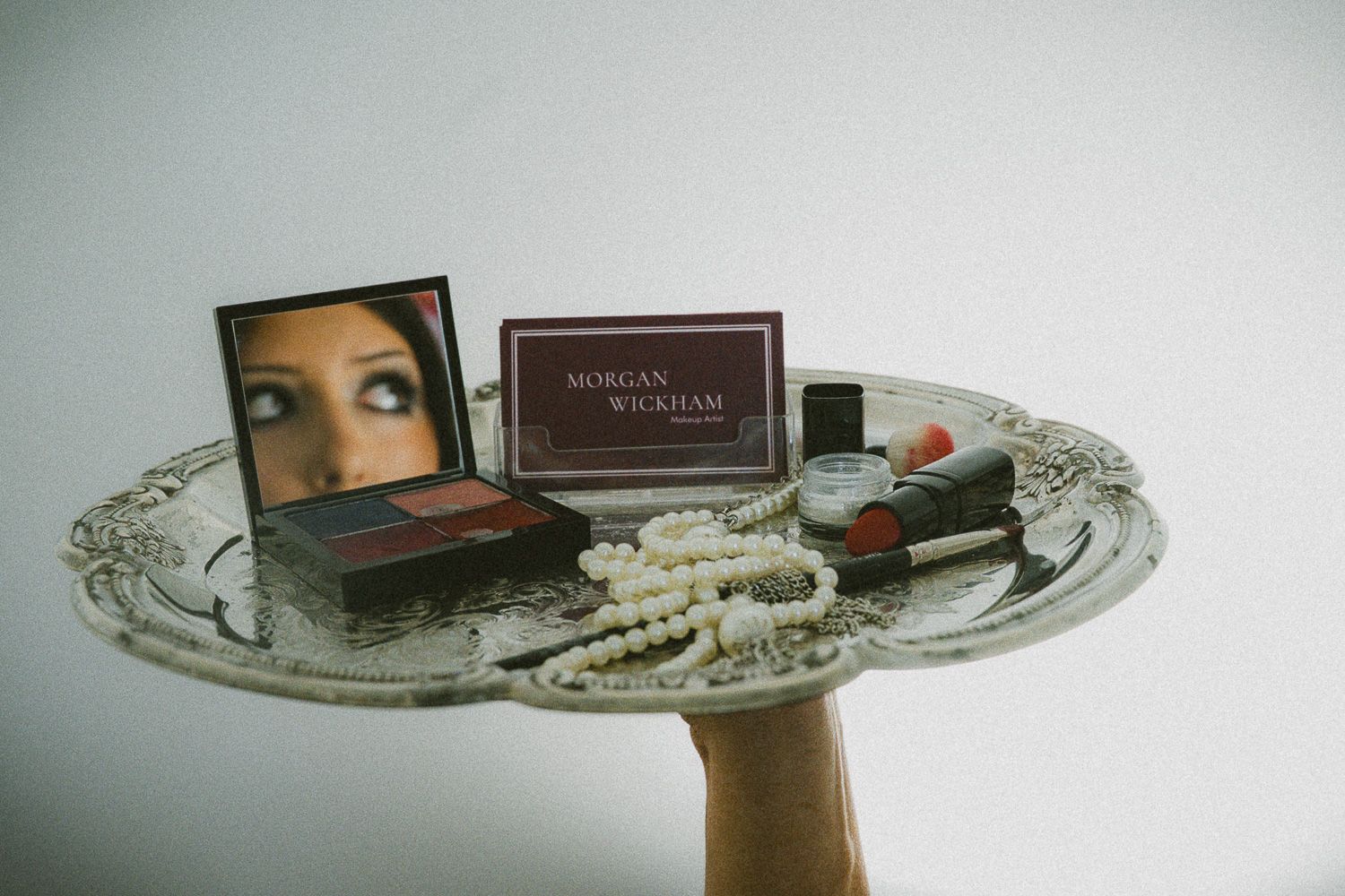 Morgan Wickham beauty makeup display with eyeshadow palette, lipstick, pearl necklace, and mirror on ornate silver tray.